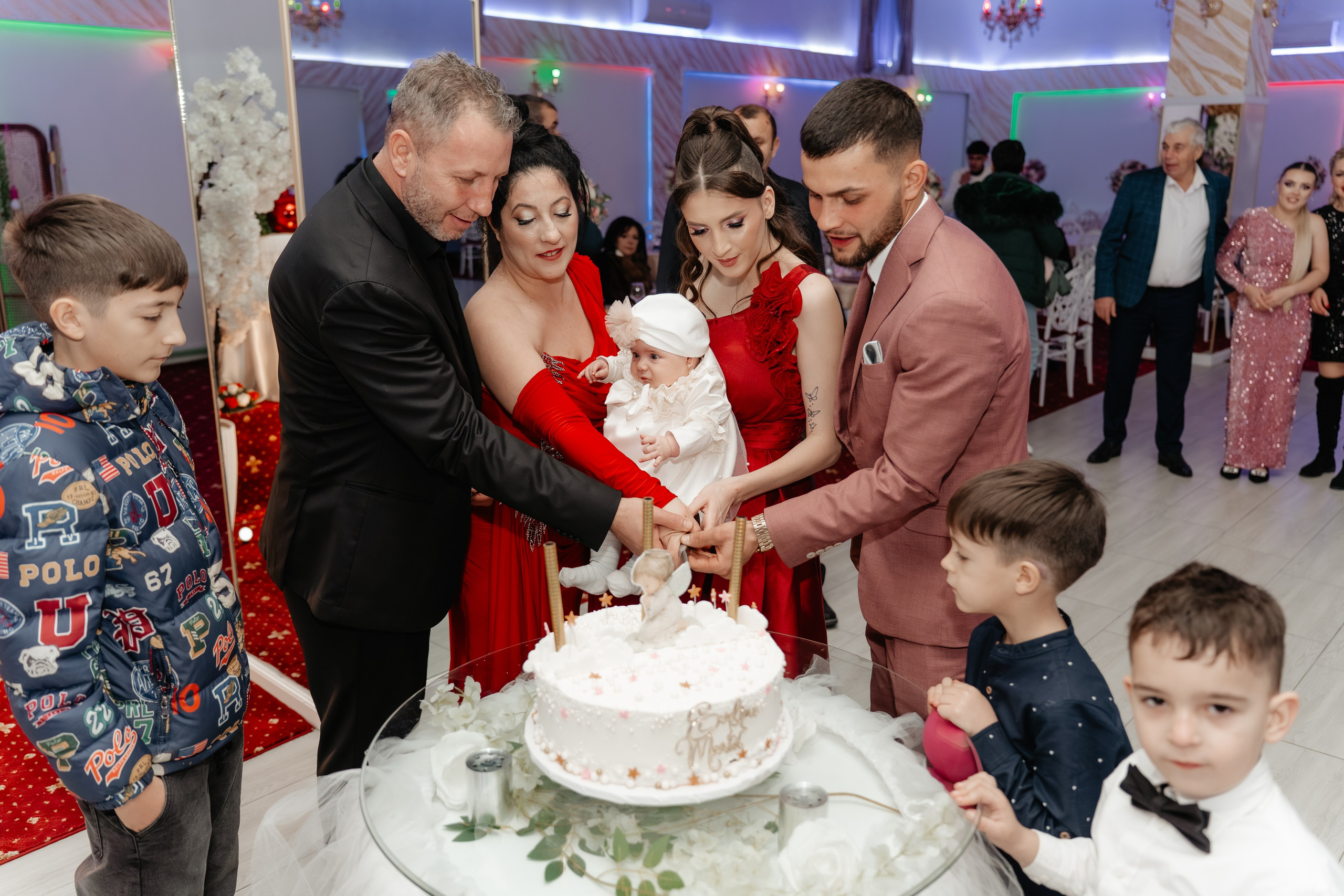 Close-up pe mâinile și fețele pline de bucurie ale părinților și nașilor în timp ce taie tortul de botez împreună cu Eveline Maria.