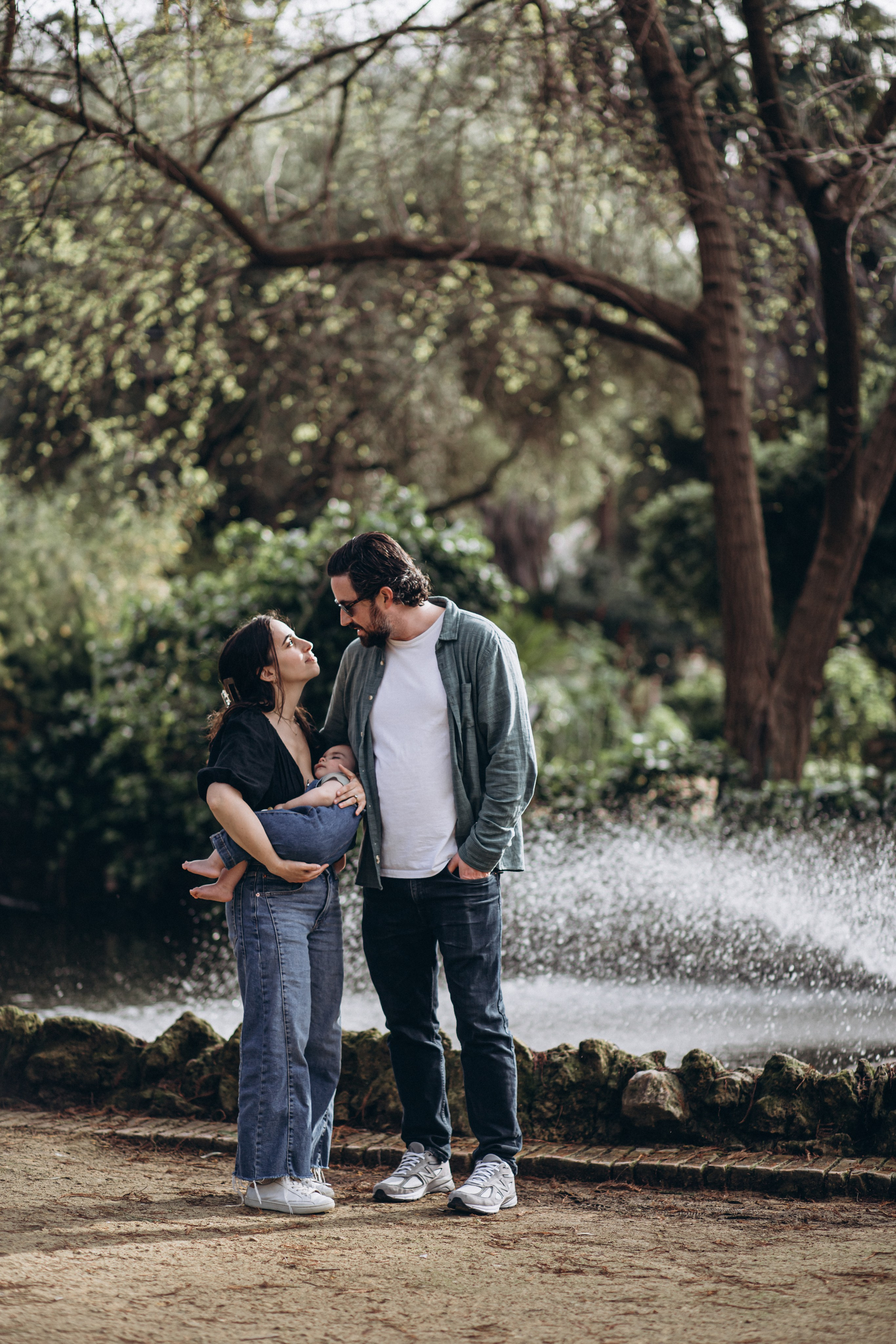 Retrato familiar amoroso en Barcelona, España — pareja sosteniendo a su bebé mientras comparten una mirada cálida, enmarcados por vegetación exuberante y la suave brisa de una fuente en un parque tranquilo.