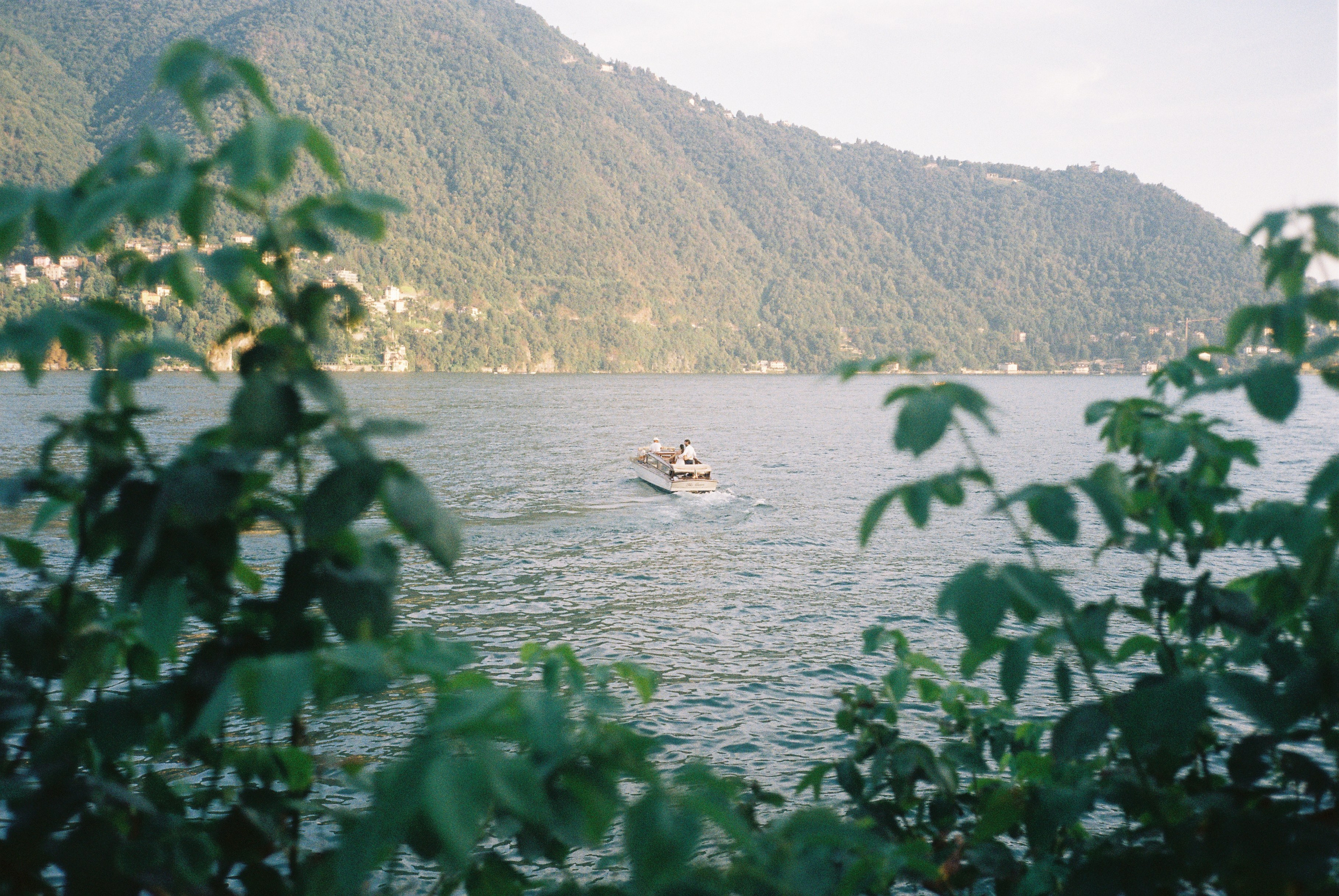Small boat on a scenic lake Como with mountains in the background, framed by foliage.