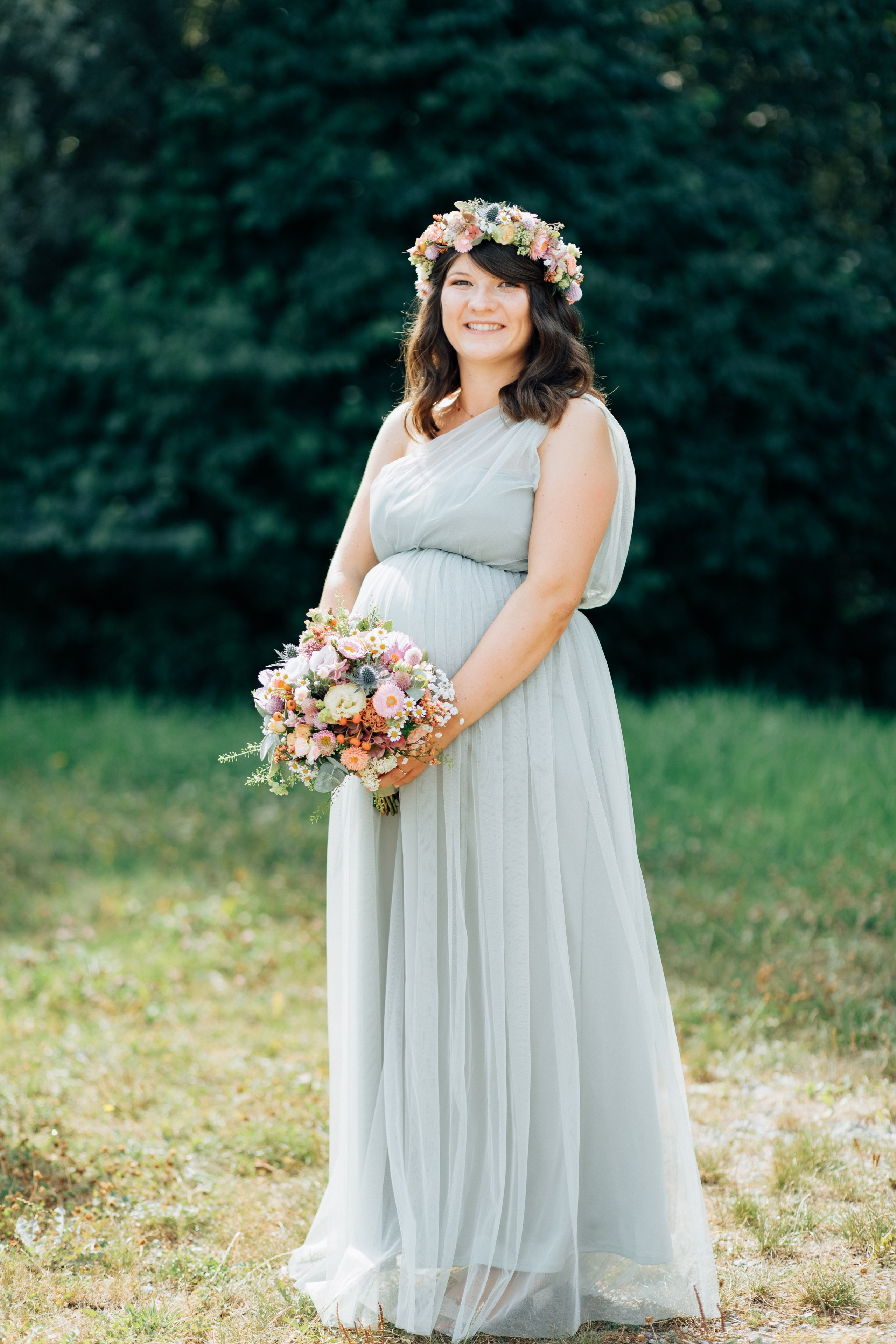Die schwangere Braut lächelt in einem wunderschönen Blumenkranz und mit einem eleganten Brautstrauß in der Hand.