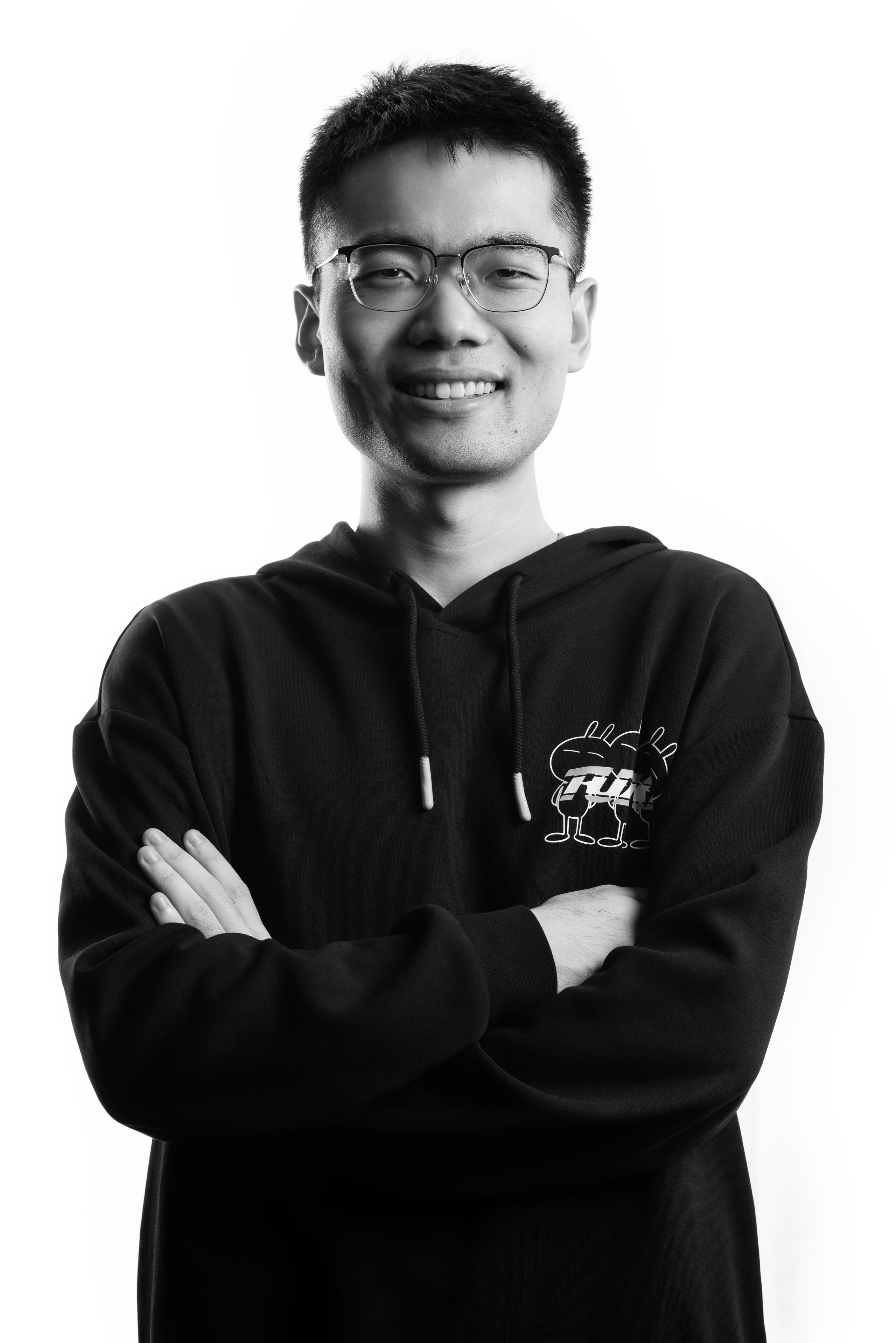 A black-and-white full-body corporate portrait of a young man with medium-length hair, wearing glasses and a dark button-up shirt with his sleeves rolled up. He stands confidently with his hands in his pockets against a bright white background, showcasing a modern and approachable style. This image highlights professional photography services for businesses in Singapore, perfect for team profiles and personal branding.