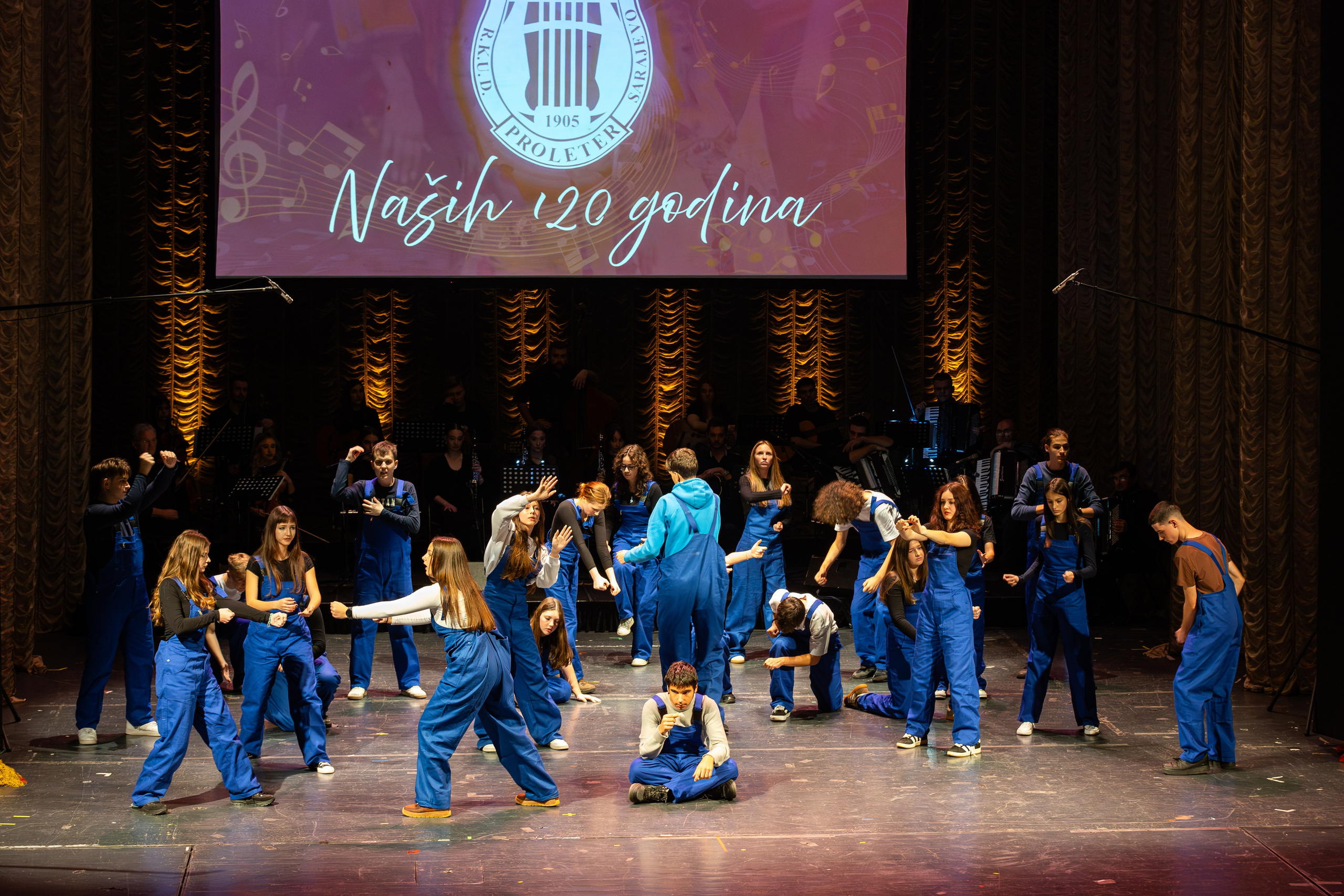 Galerija fotografija sa svečanog koncerta RKUD Proleter u Narodnom pozorištu Sarajevo, prikazuje nastupe izvođača, svečanu atmosferu, bogatu scenografiju i publiku u prepunoj dvorani.