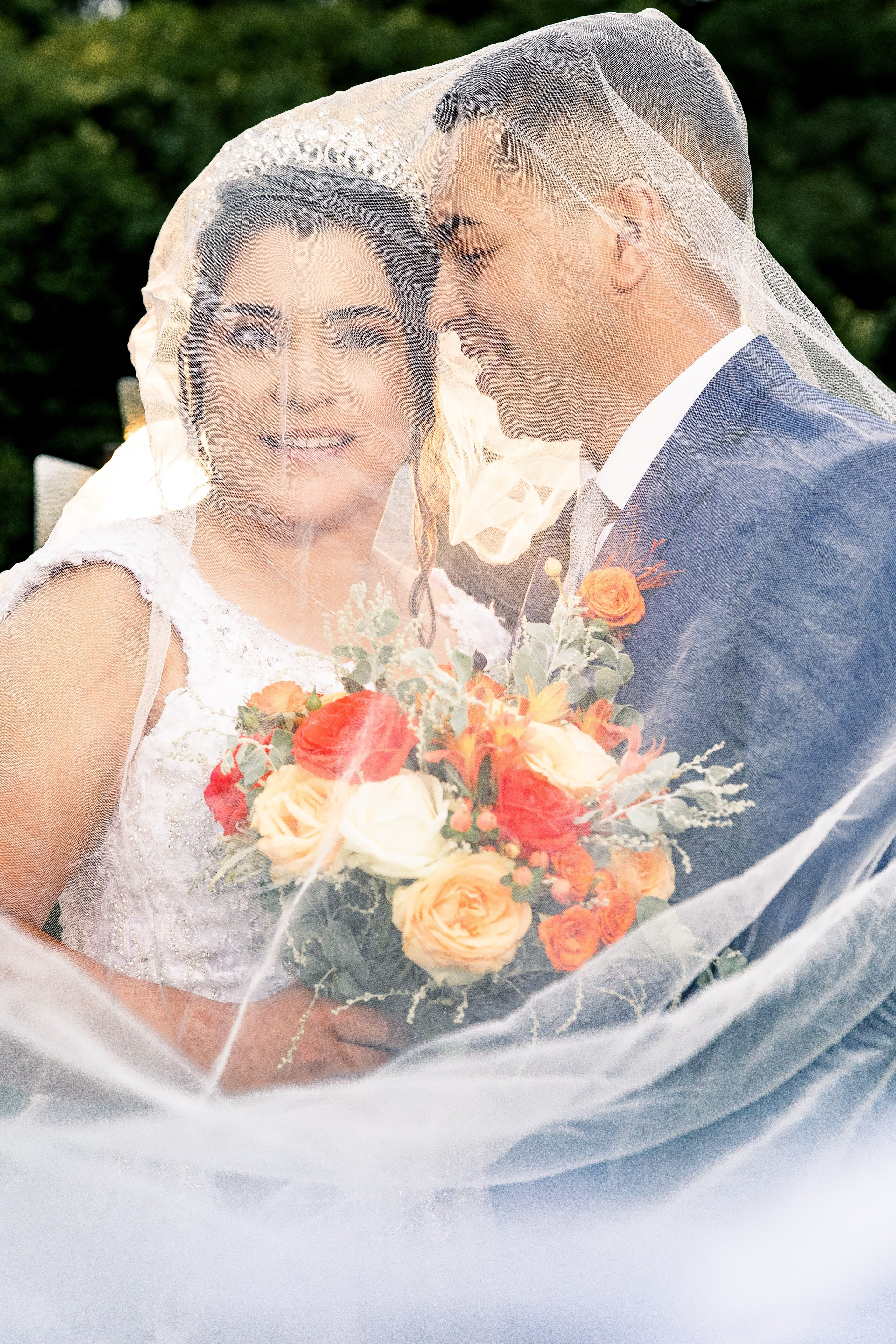 Noiva com olhar direto e buquê vibrante, noivo sorrindo ao seu lado sob o véu transparente.