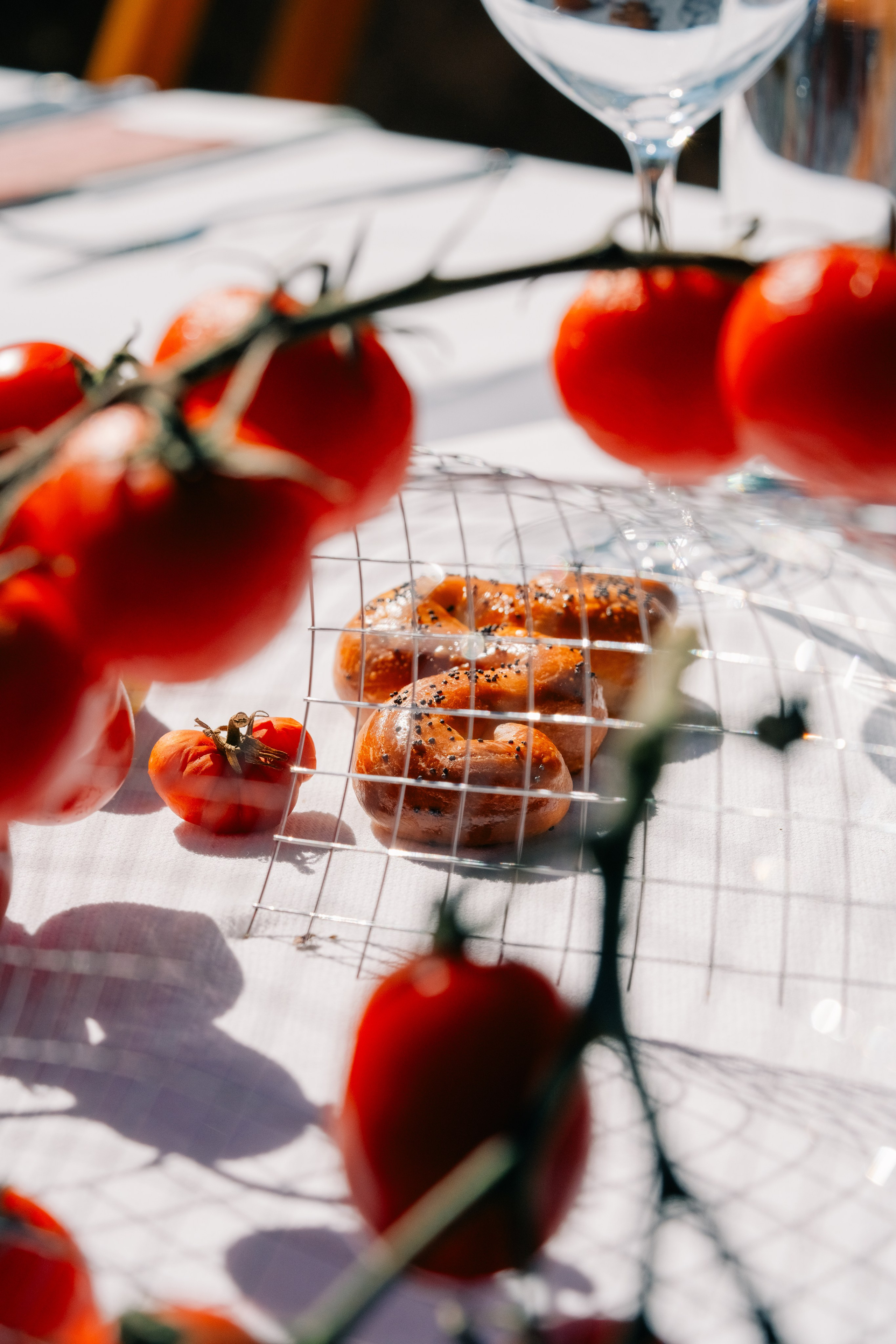 Крупный план свежих помидоров и кренделя на залитом солнцем столе на открытом воздухе. Художественный крупный план свежих помидоров на ветке и кренделя на залитом солнцем обеденном столе на открытом воздухе, создающий яркую и аппетитную сцену.