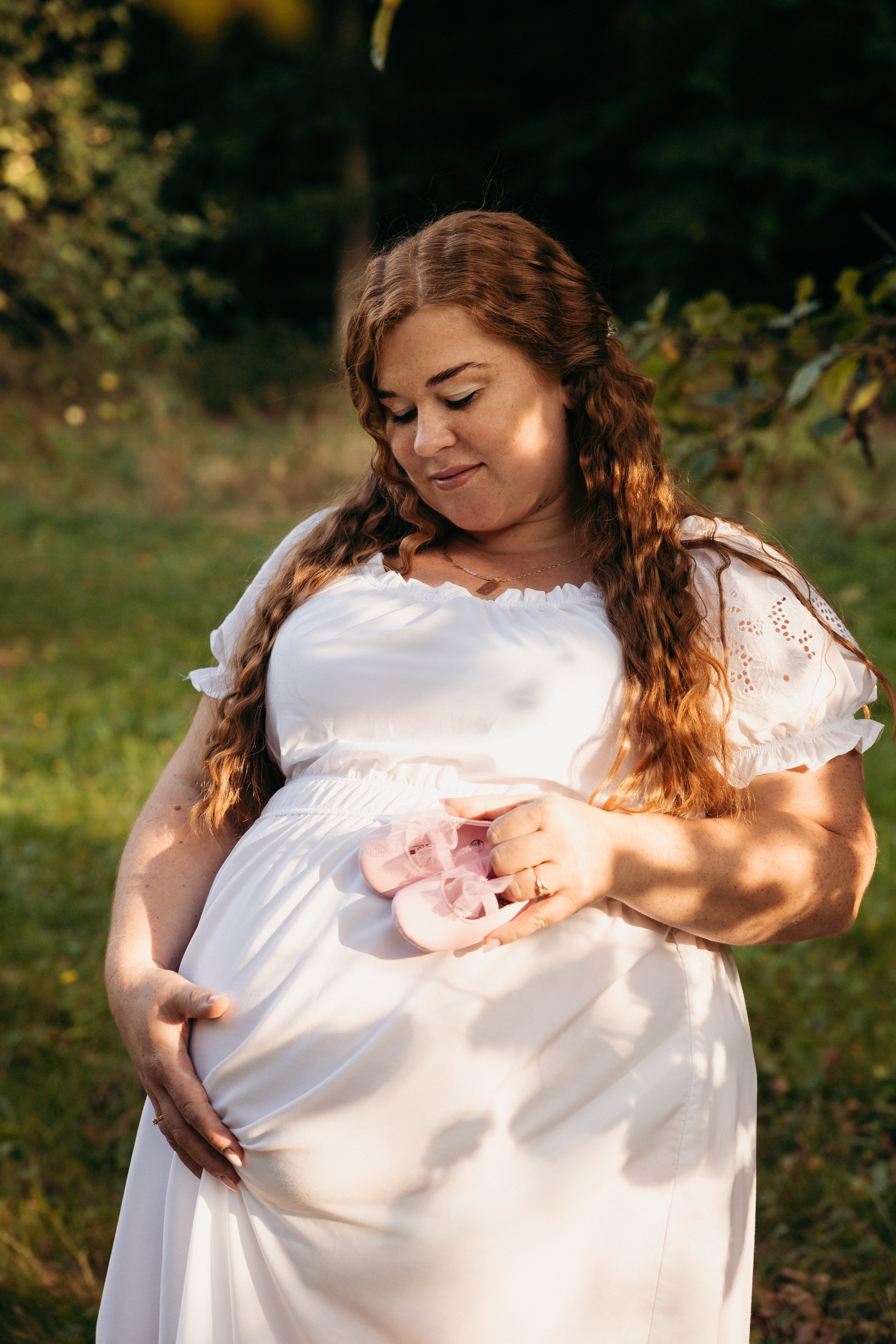 Těhotná žena v bílých šatech stojí v přírodě – těhotenské focení v exteriéru 