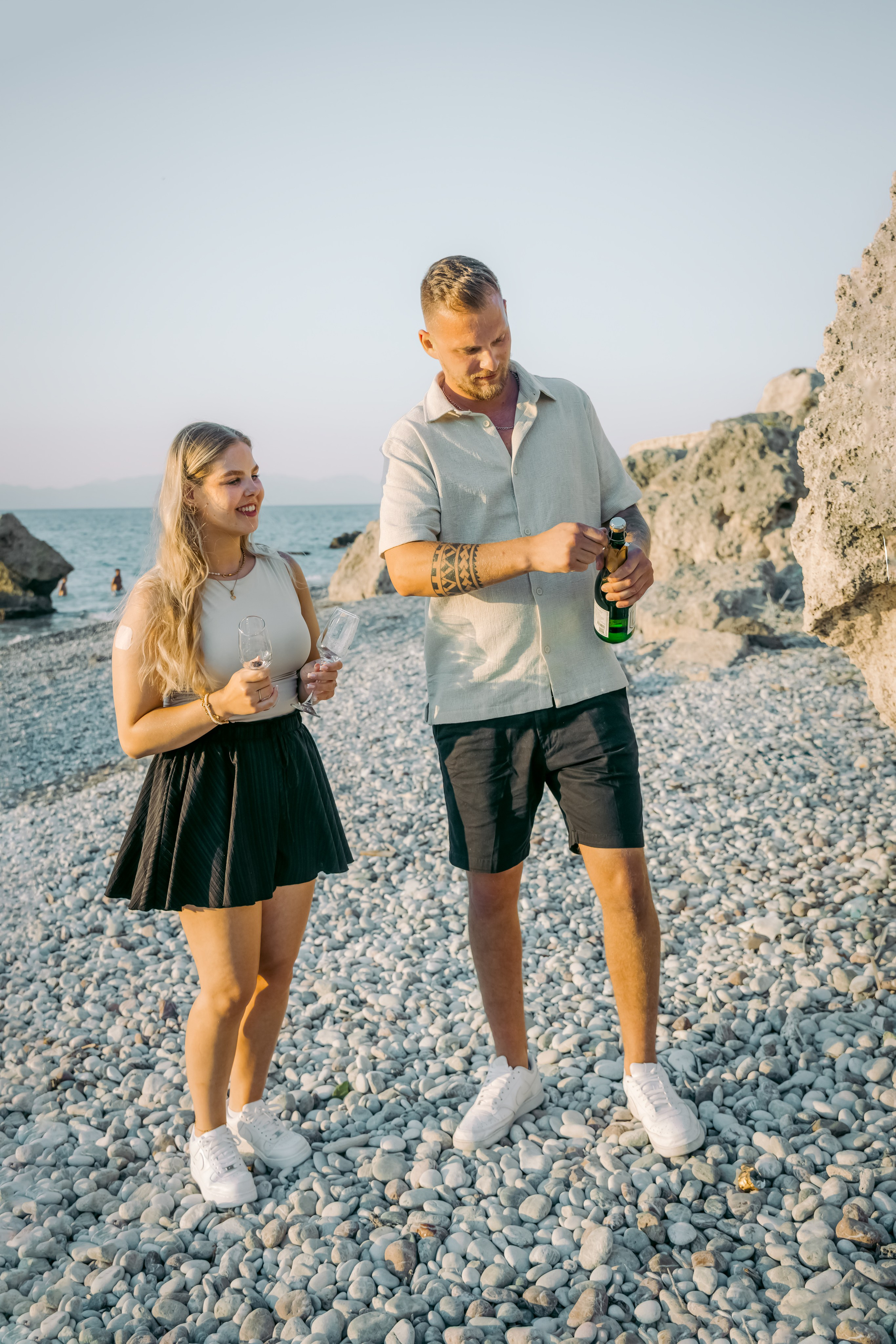 "Ein Paar spaziert Hand in Hand am steinigen Ufer von Kato Petres auf Rhodos entlang. Sie gehen direkt am Wasser, während im Hintergrund die hohen, zerklüfteten Kalksteinfelsen aufragen und das blaue Meer sanft gegen die Kiesel brandet."