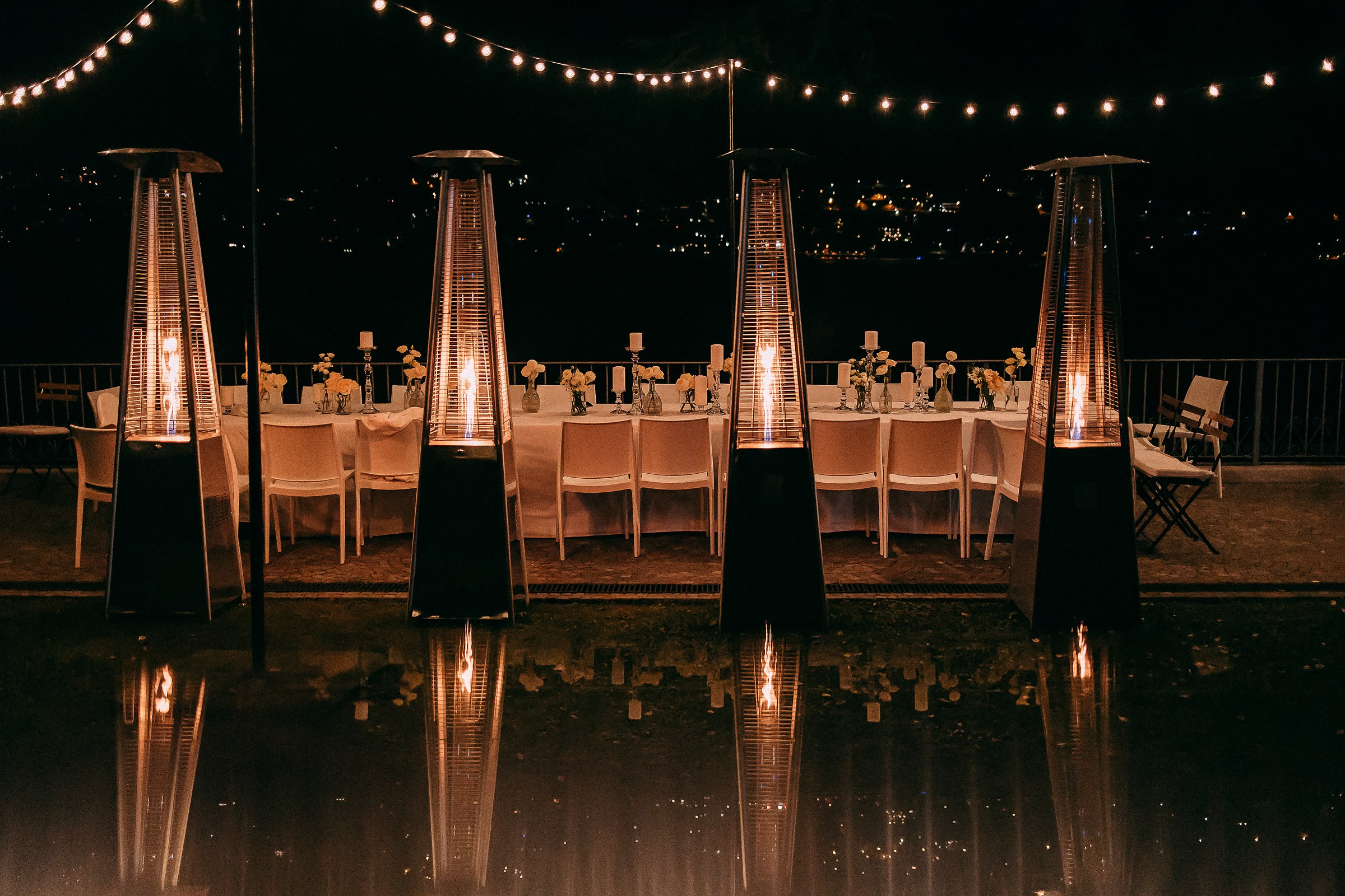 Night view of an elegantly set dinner table illuminated by string lights, with heaters reflecting in the water.