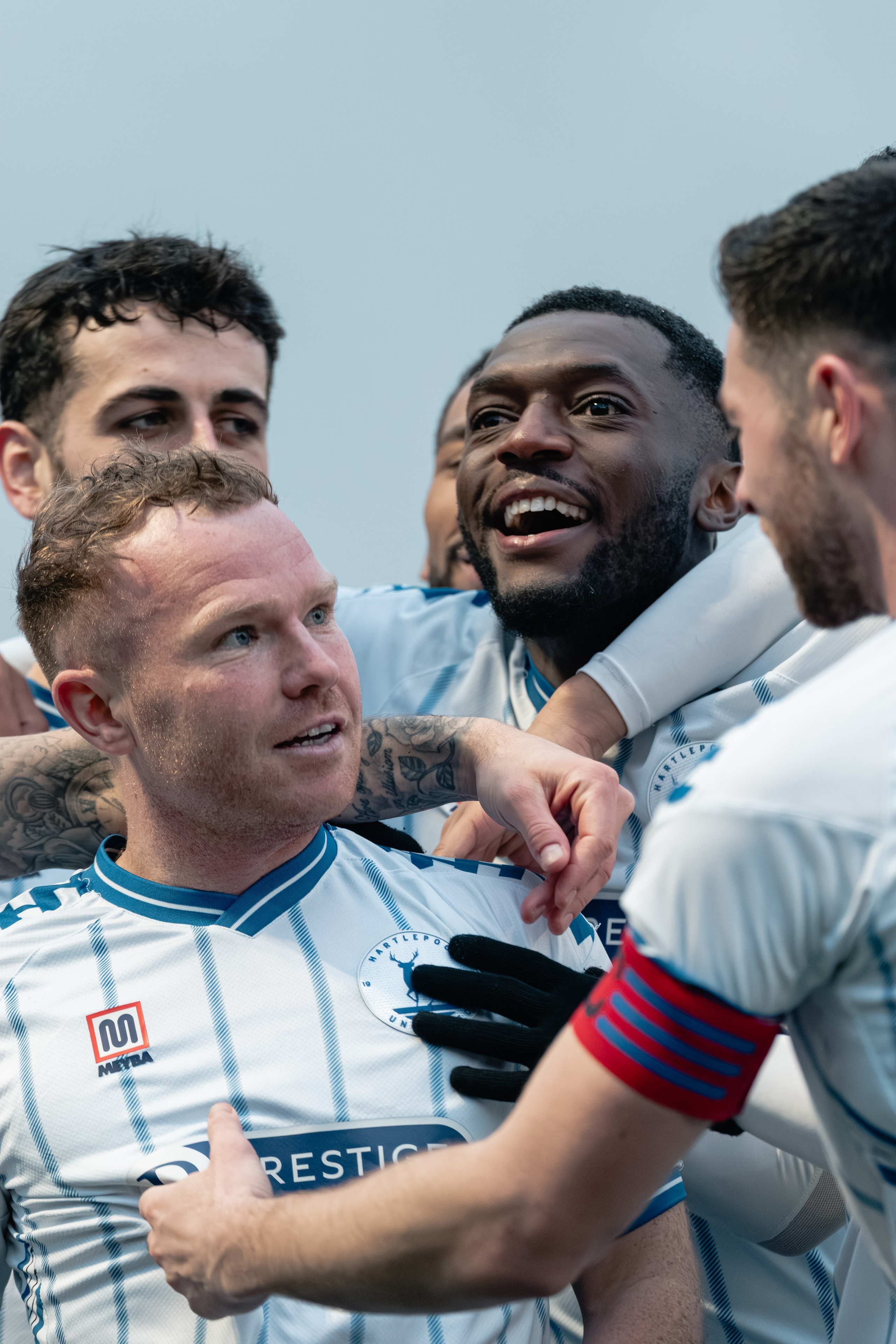 Close-up of Hartlepool United F.C. players in white-and-blue kits celebrating together in a huddle, smiling and embracing after a goal.