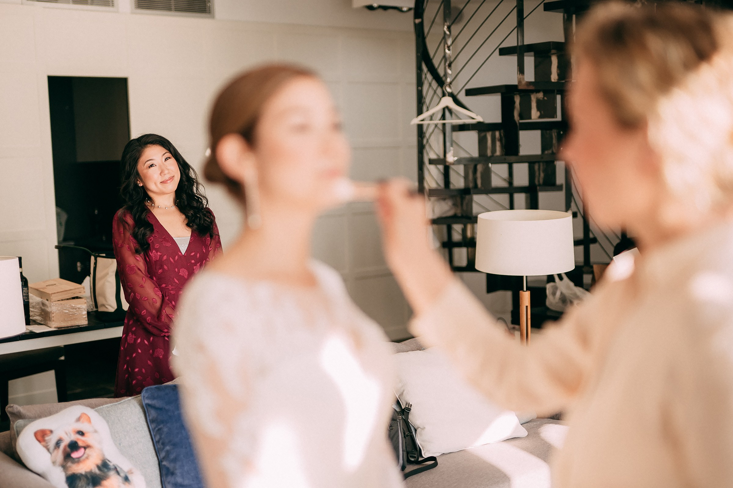Bridesmaid watches the bride’s makeup being done, smiling in the background of a cozy room.