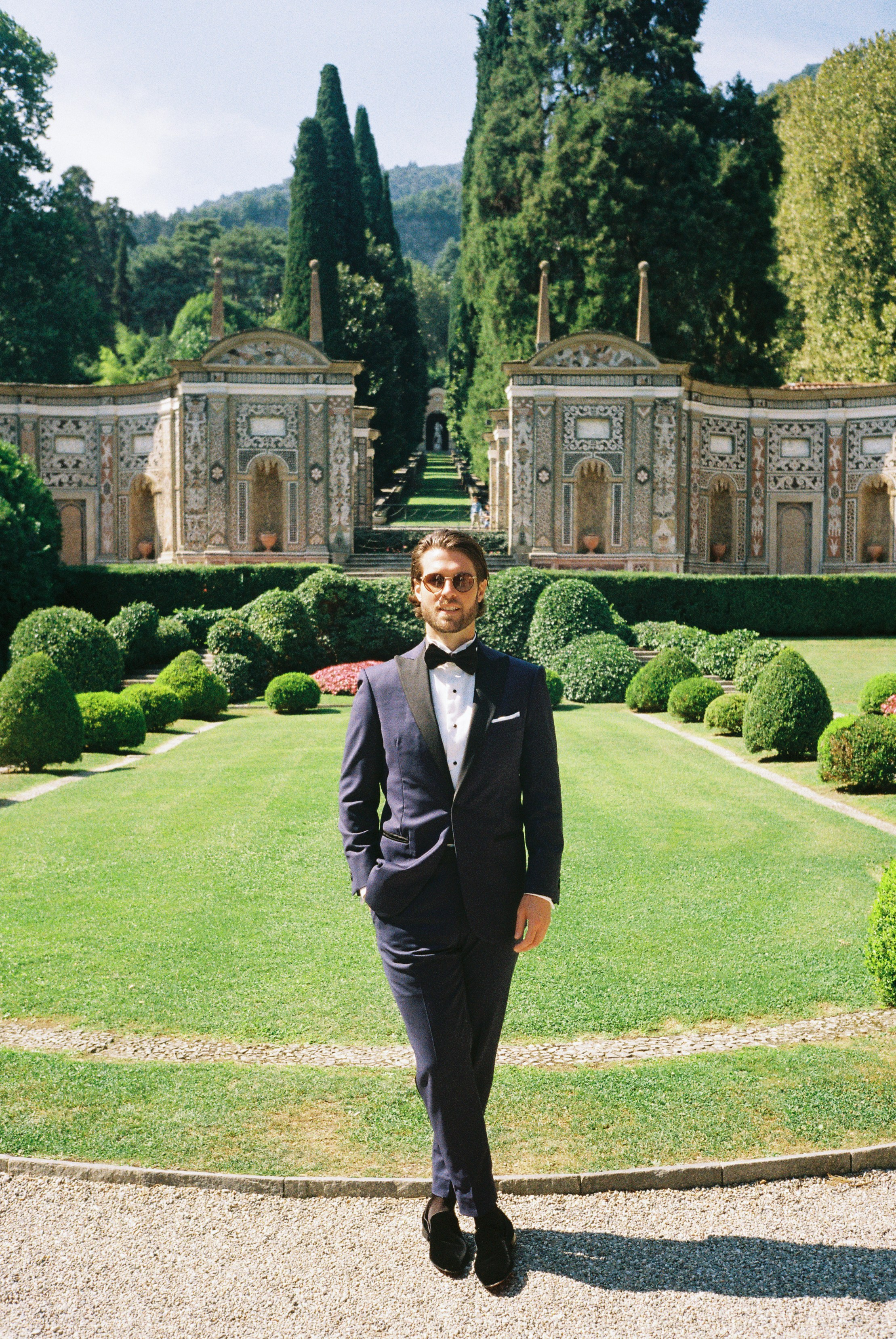 Groom in dark tuxedo stands confidently in landscaped garden, framed by ornate stone walls.