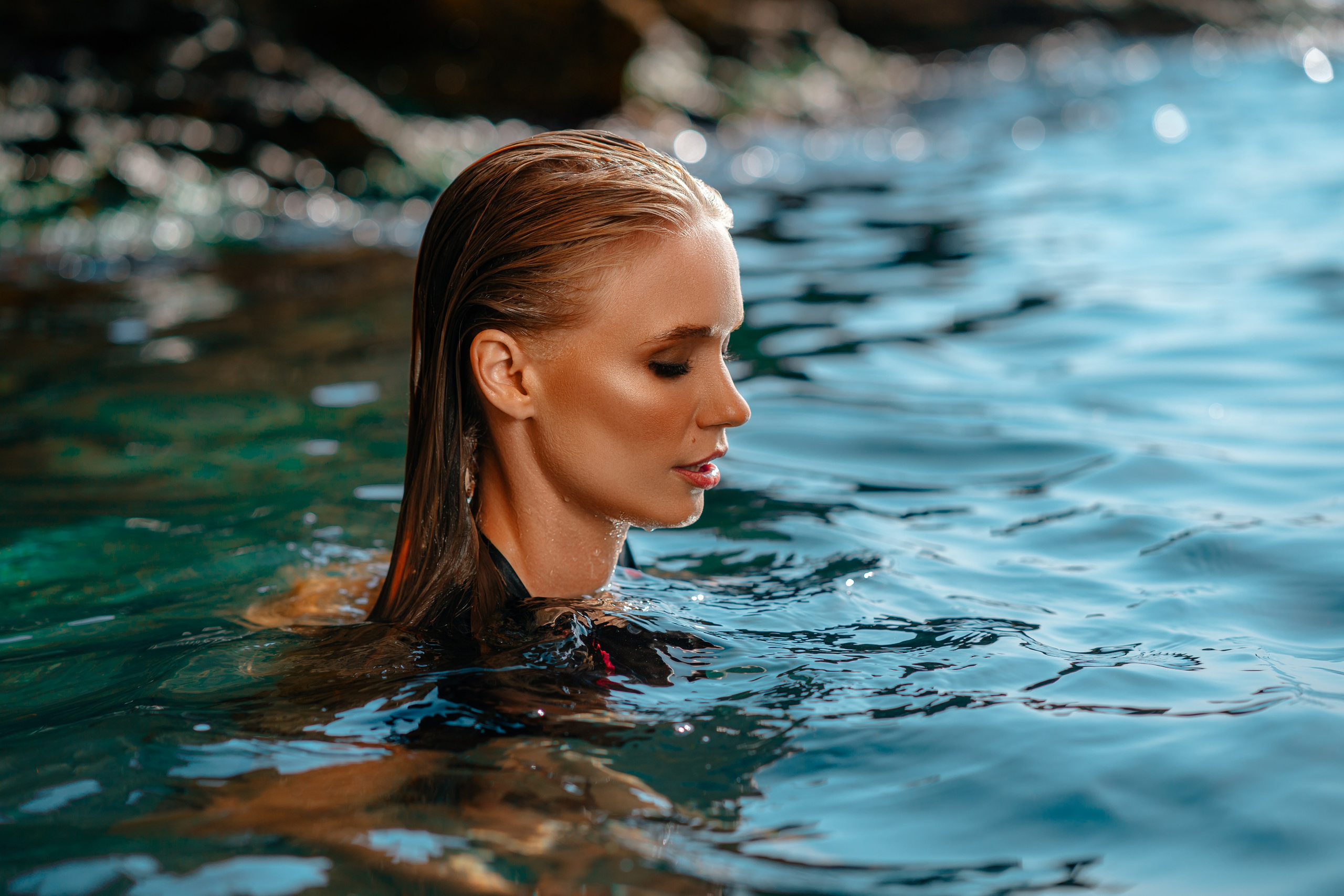 An intimate profile portrait of a young woman immersed in the sea, her wet hair shimmering in the warm embrace of sunlight. The play of sunlight on her skin reveals a captivating dance of light and shadow, accentuating the delicate contours of her face. The calm sea serves as a reflective canvas, mirroring the tranquility and grace of the moment, creating a serene atmosphere that beautifully complements her captivating presence