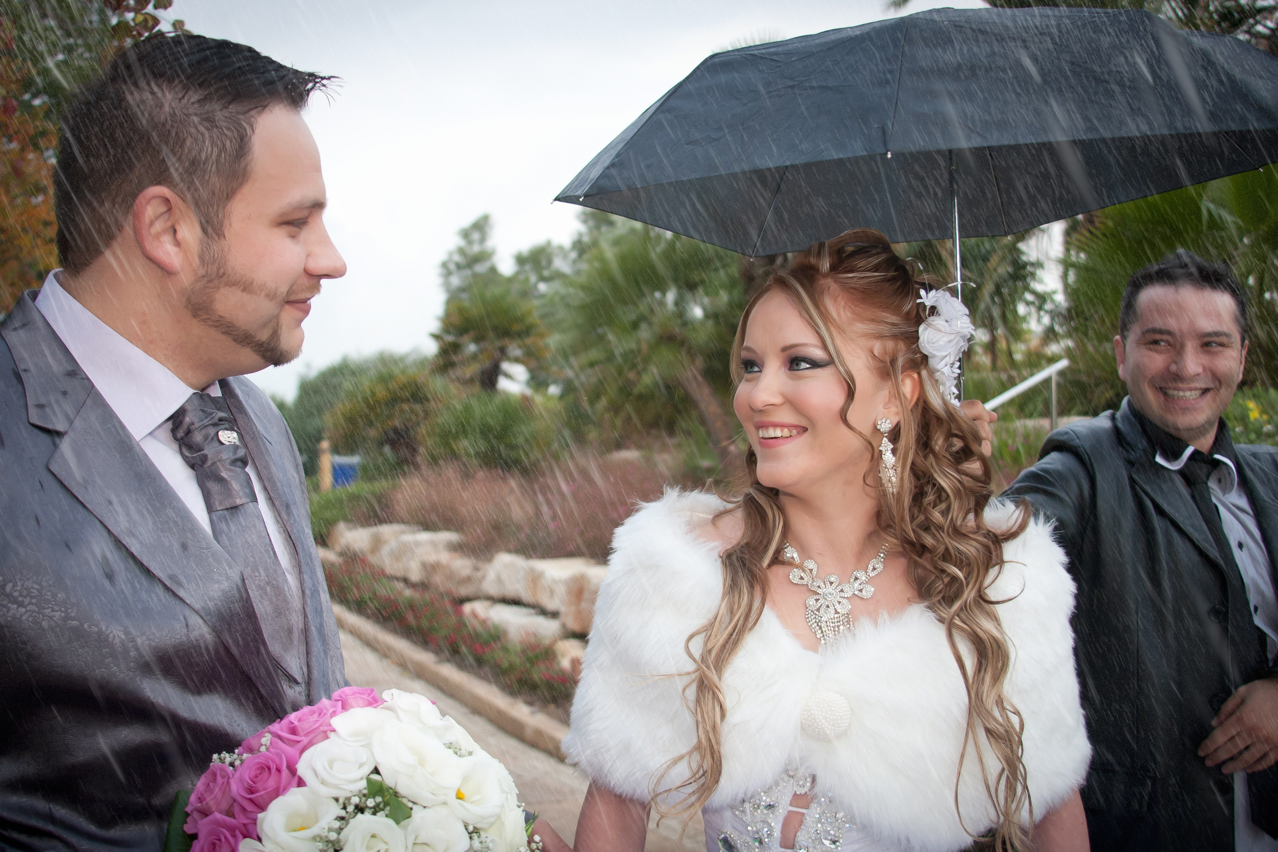 A groom and bride smile at each other under the rain, sharing a romantic moment. The groom holds a bouquet of white and pink roses, while a man behind them holds an umbrella, laughing. The bride wears a white fur stole and a sparkling necklace, her hair adorned with a floral accessory. The scene captures the joy and spontaneity of a rainy wedding day.