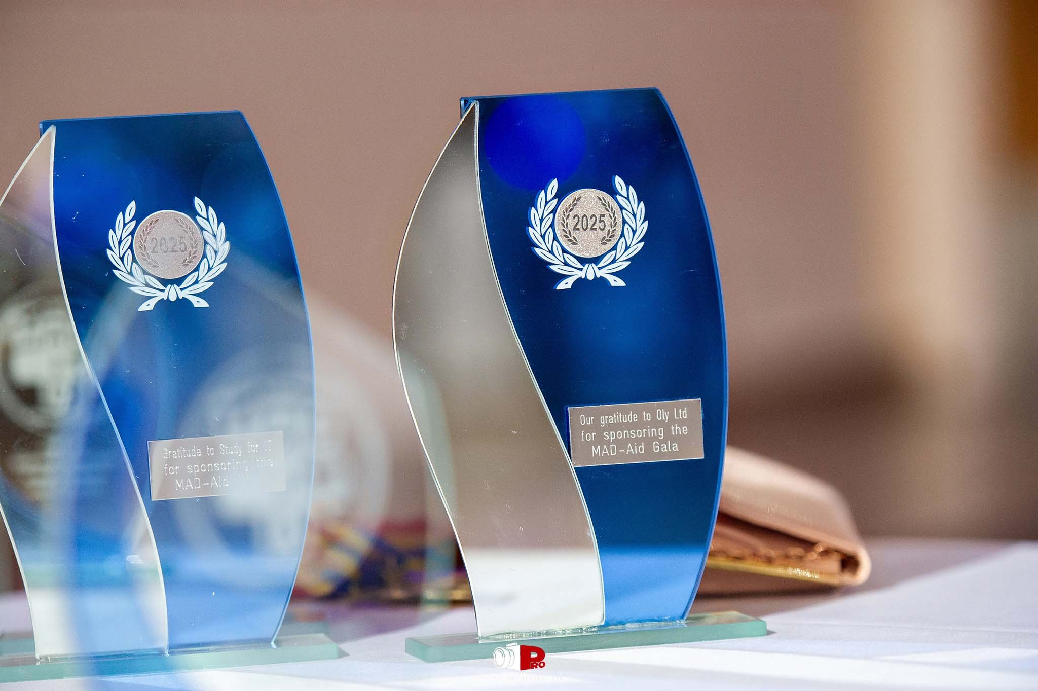 Two striking blue awards with "MAD-Aid Gala 2025" engraved on them are displayed on a table.