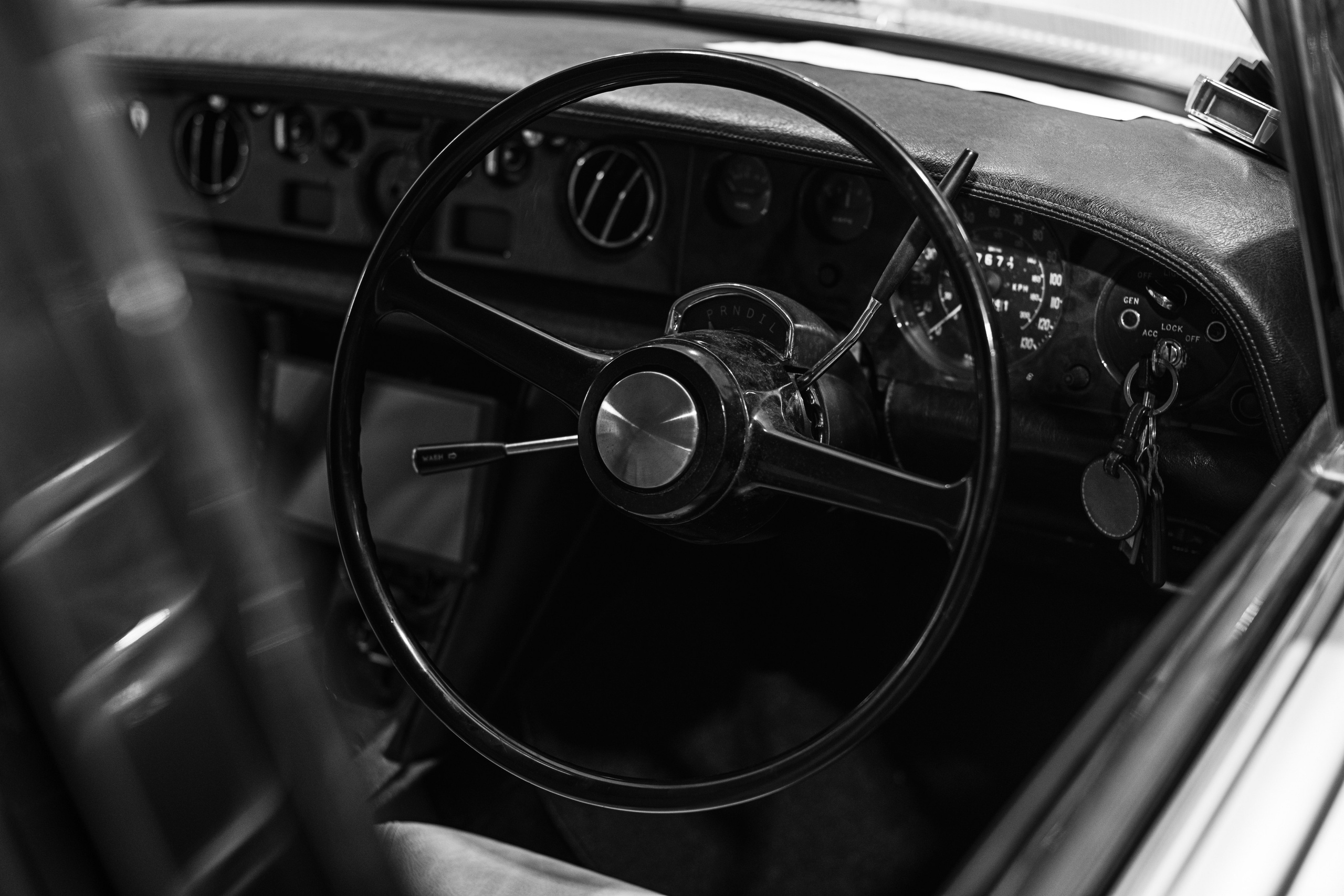 Black-and-white close-up photograph of the steering wheel and dashboard of a classic Rolls-Royce, highlighting the vintage instrument panel and intricate design elements. Captured by Mirrorklezz Photography at The Garage by Classic Motorworks in Singapore, this image showcases the timeless elegance and precision craftsmanship of luxury automotive interiors. https://www.classicmotorworks.com.sg/