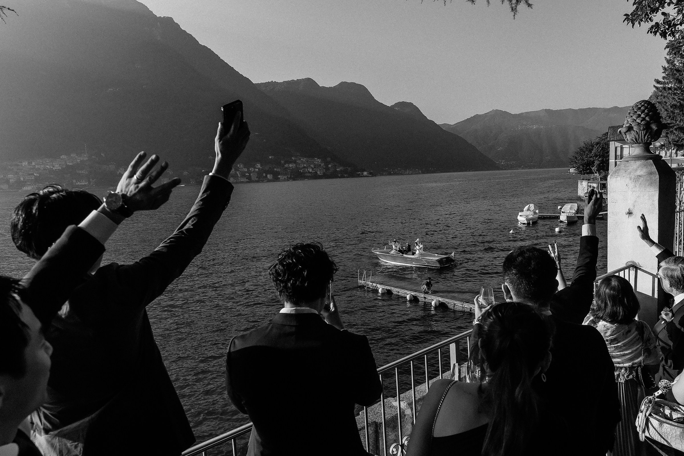 Guests waving and capturing a boat with the bride and groom as it departs, set against scenic mountains and lake.