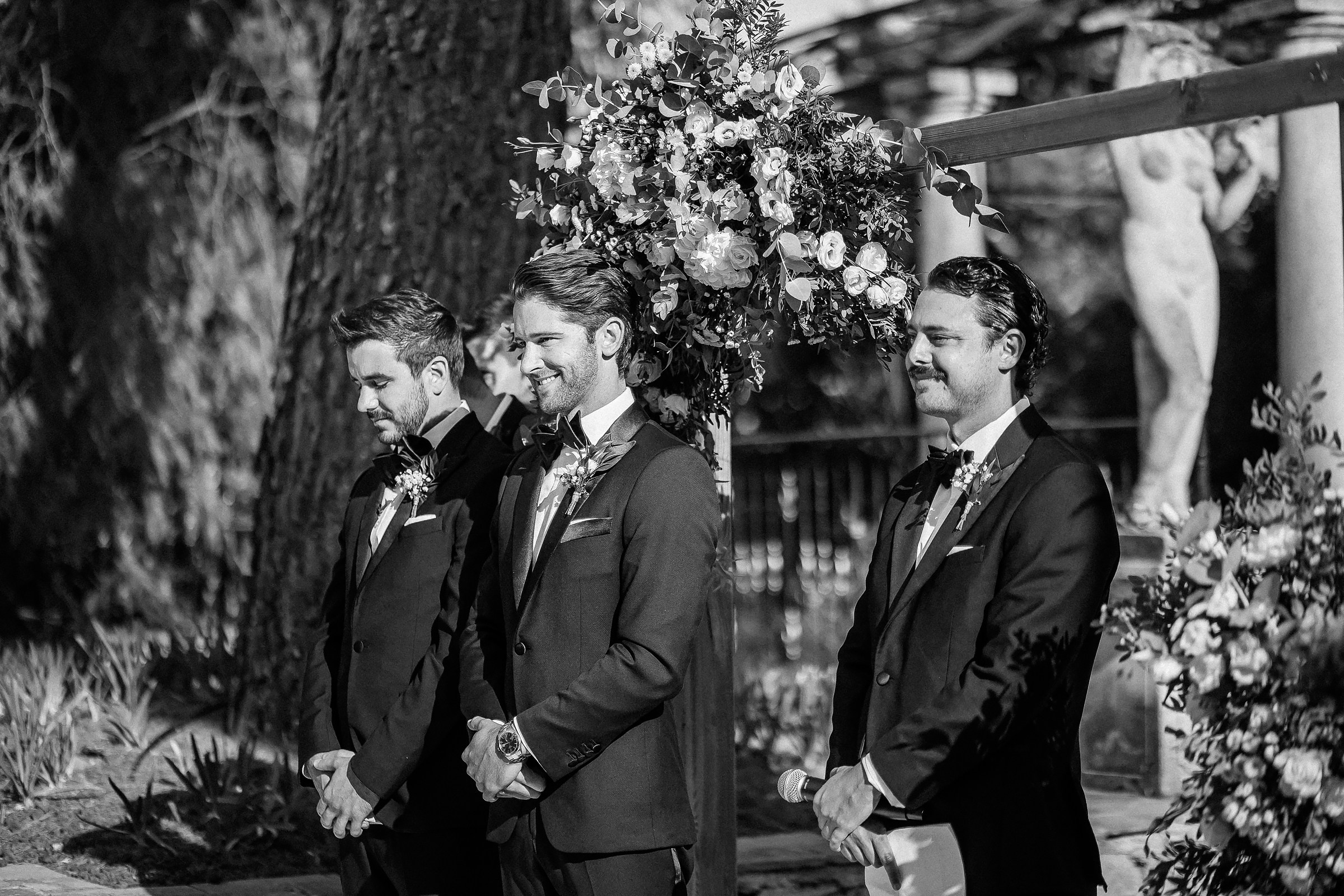 The groom and his groomsmen stand together under a floral arch, exuding anticipation and camaraderie in the sunlight during the wedding ceremony. 