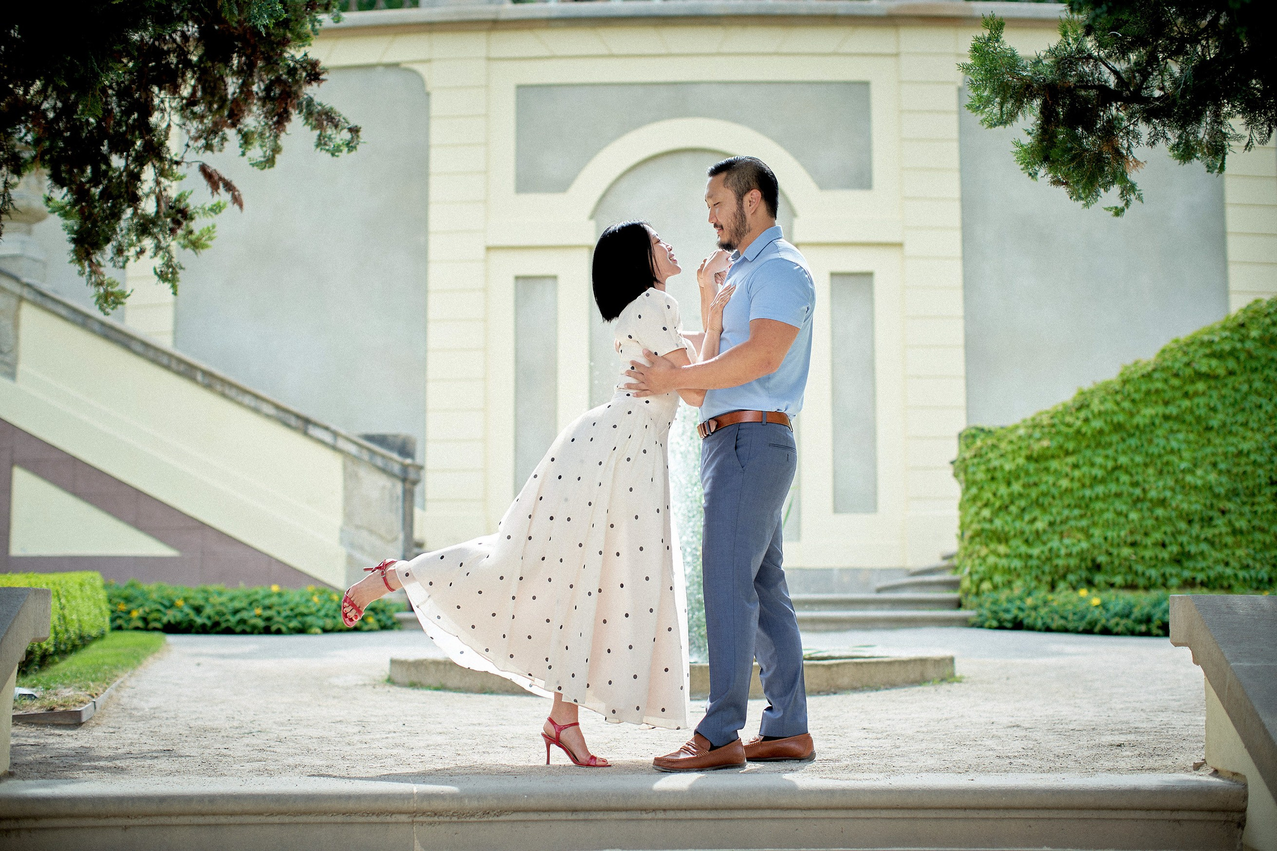 Playful woman grasping her partner's collar and lifting her red heel in Vrtba Garden engagement session.