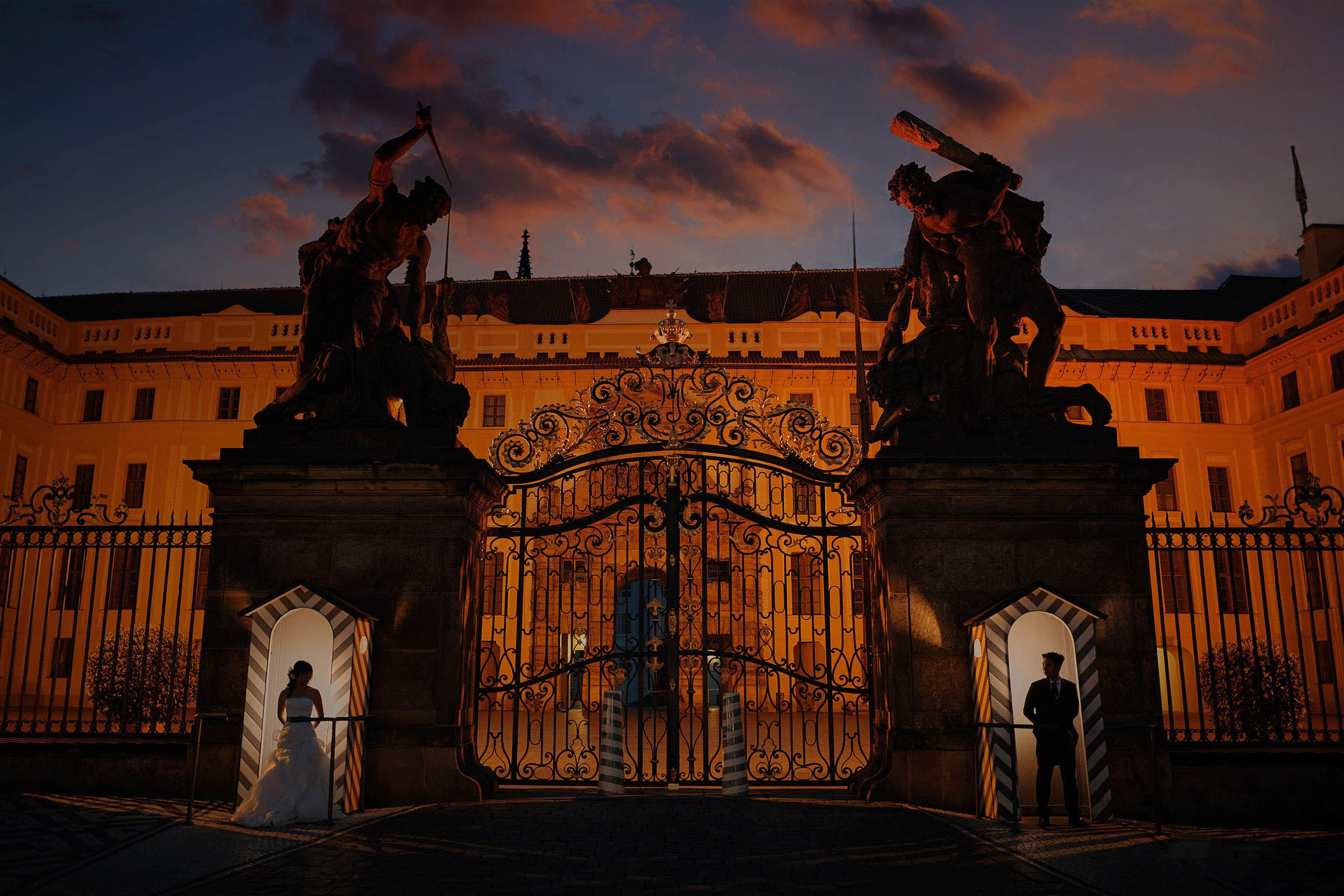 Stylish Asian bride and groom stand separately in the guardshacks of Prague Castle at night as the sky flares above