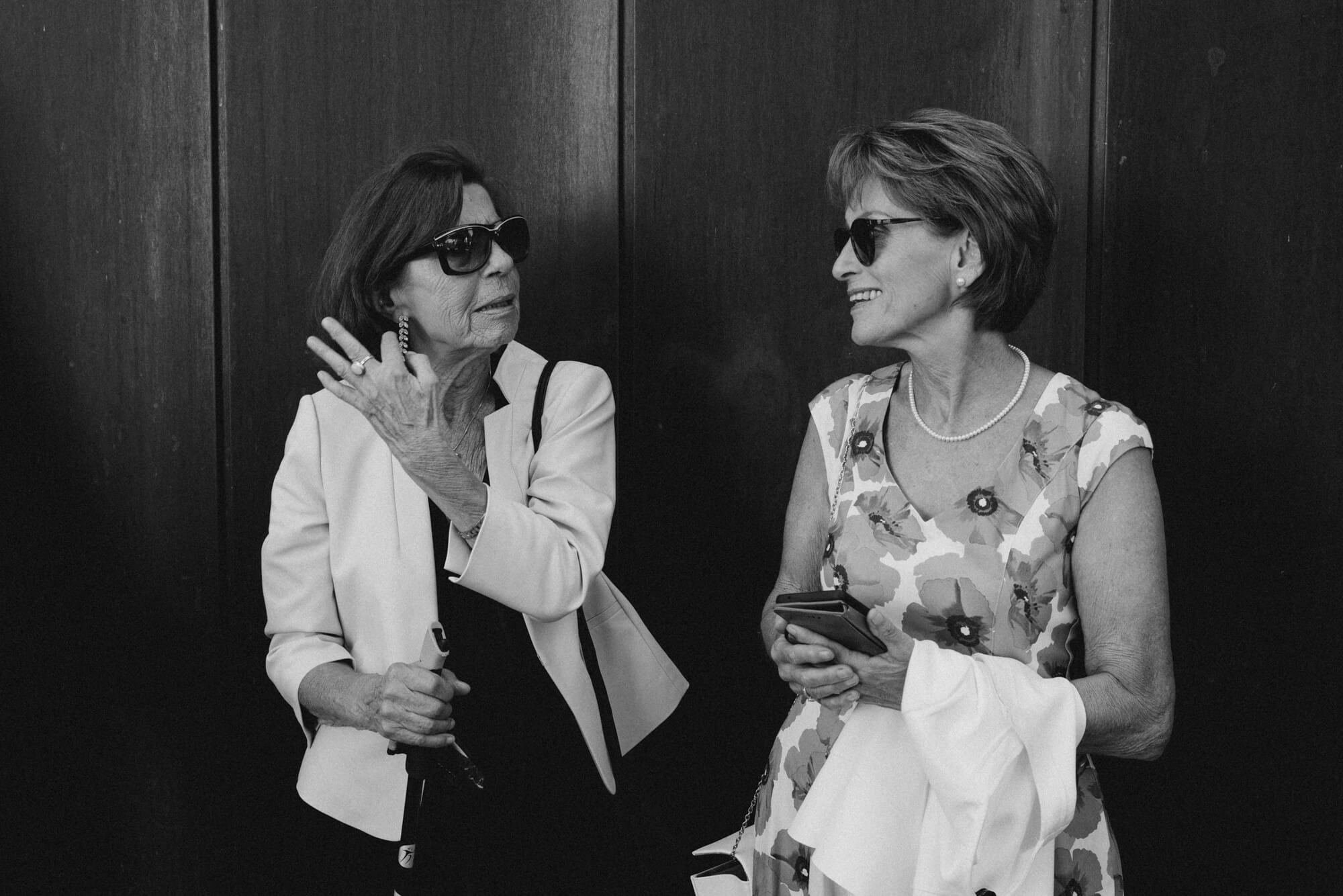 Two women in elegant attire talking outside the venue before the ceremony, one dabbing her eye; black and white
