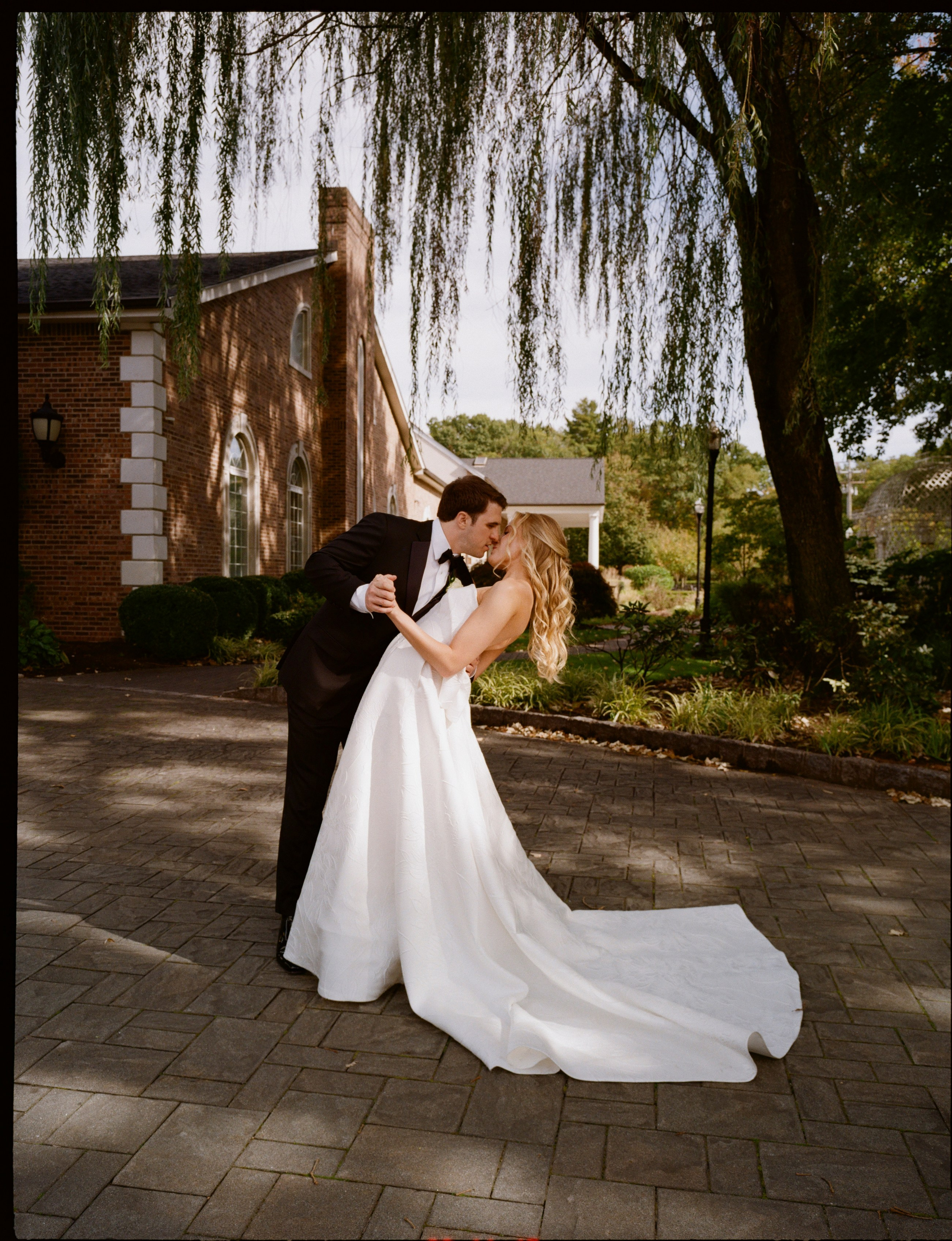 Romantic dip kiss with Emily’s wedding dress flowing across the stone pathway