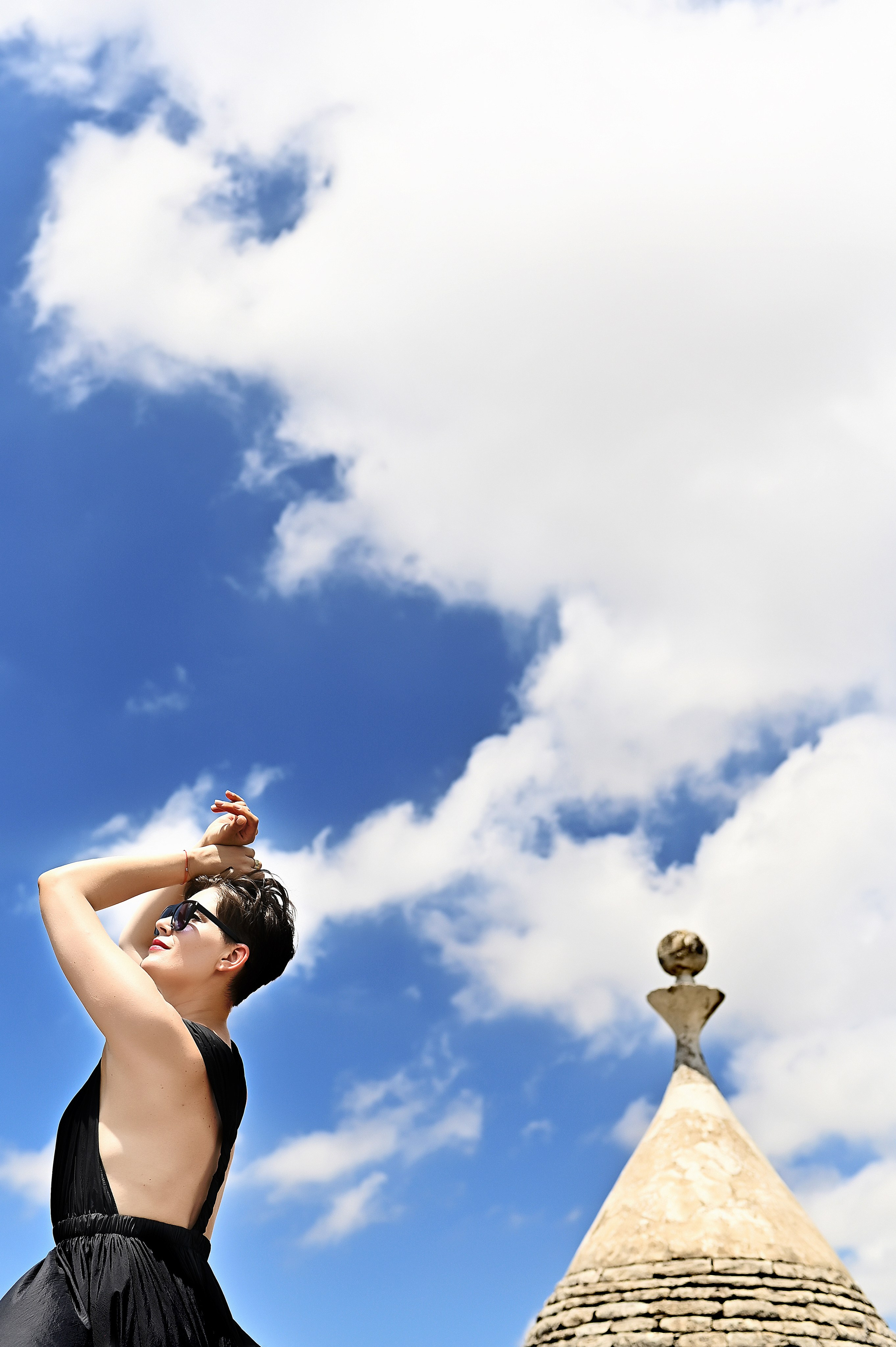 Woman in a black dress raises her arms to the sky, standing near a trullo roof under blue sky and clouds.