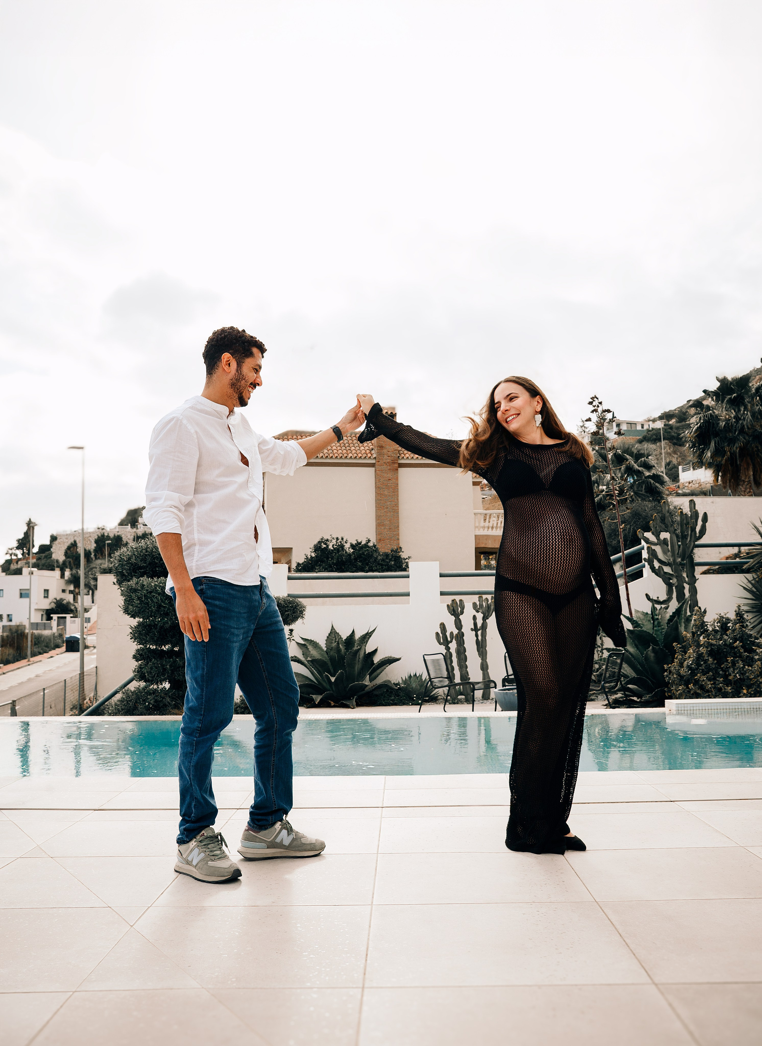 Alegre retrato de embarazo en Valencia, España — pareja bailando junto a una moderna terraza con piscina; la futura madre radiante con vestido negro de malla y el padre sonriente.