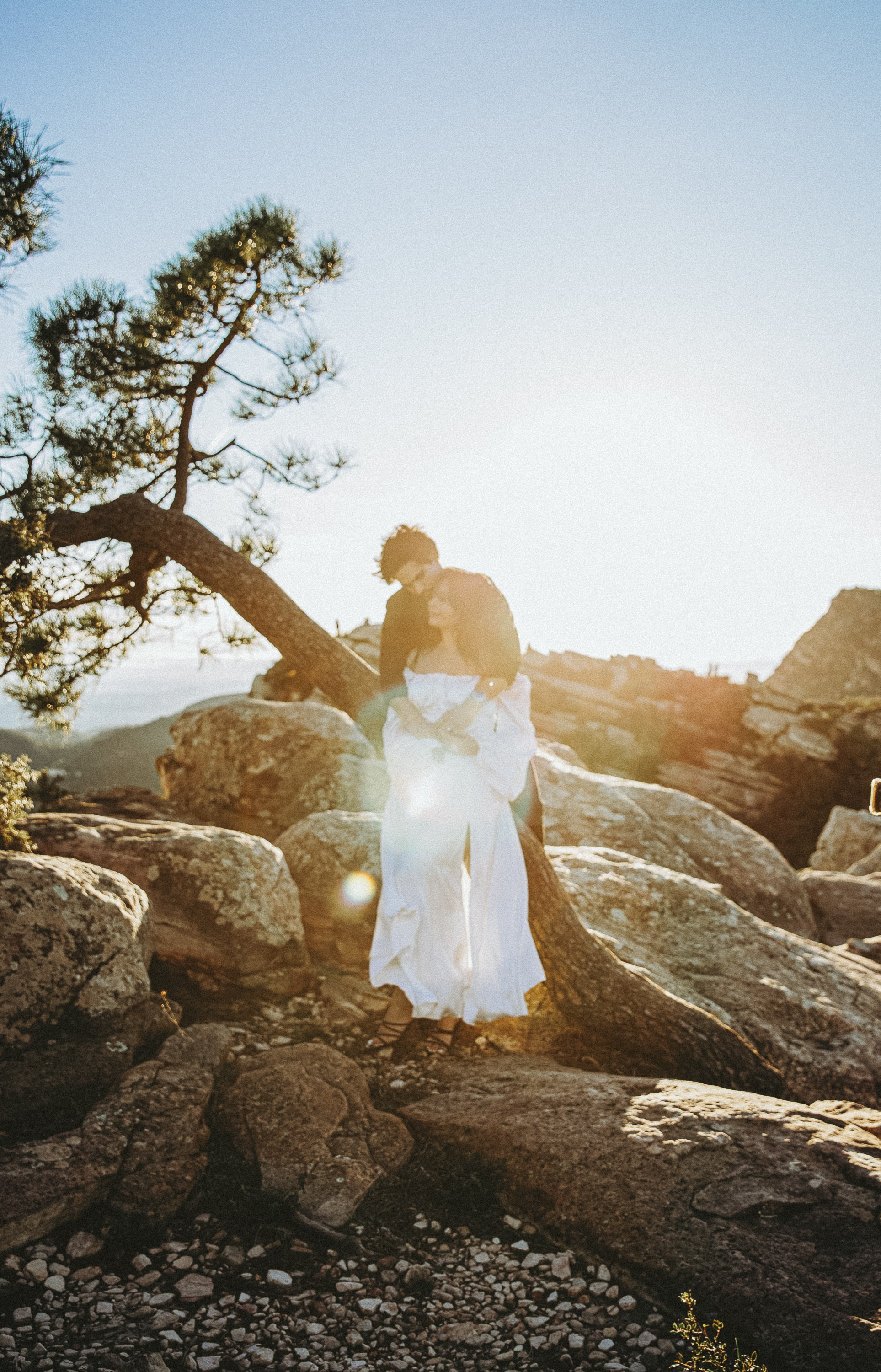 Engagement photoshoot in València, Spain, featuring a romantic couple embracing among rocky hills and pine trees at golden hour, with soft sunlight and a flowing white dress creating a cinematic, emotional mood — a timeless wedding love story image ideal for engagement photography, pre-wedding sessions, elopements, and professional wedding photoshoots in València and across Spain.