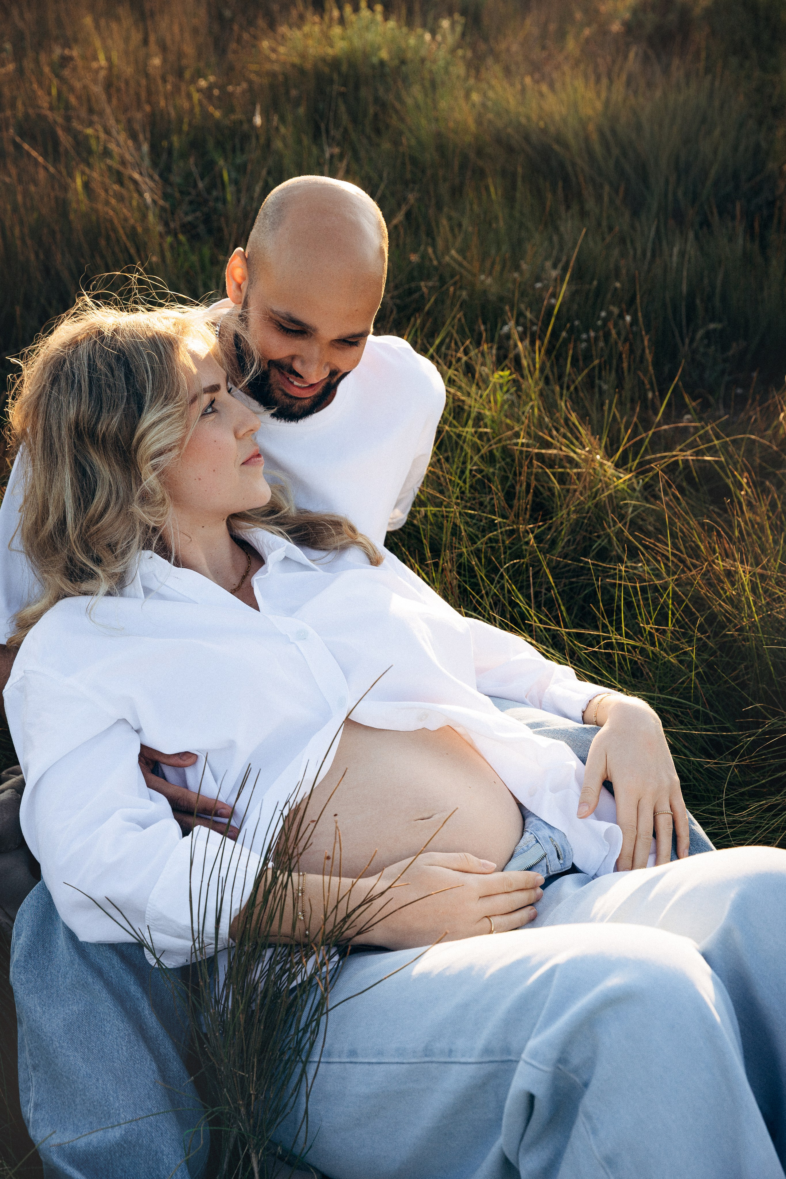 Mujer embarazada con un vestido blanco fluido posando junto al mar en Valencia, España, sosteniendo suavemente su barriga durante una sesión natural de embarazo al aire libre con suave luz de atardecer y fondo costero mediterráneo, capturando un momento elegante y emocional.