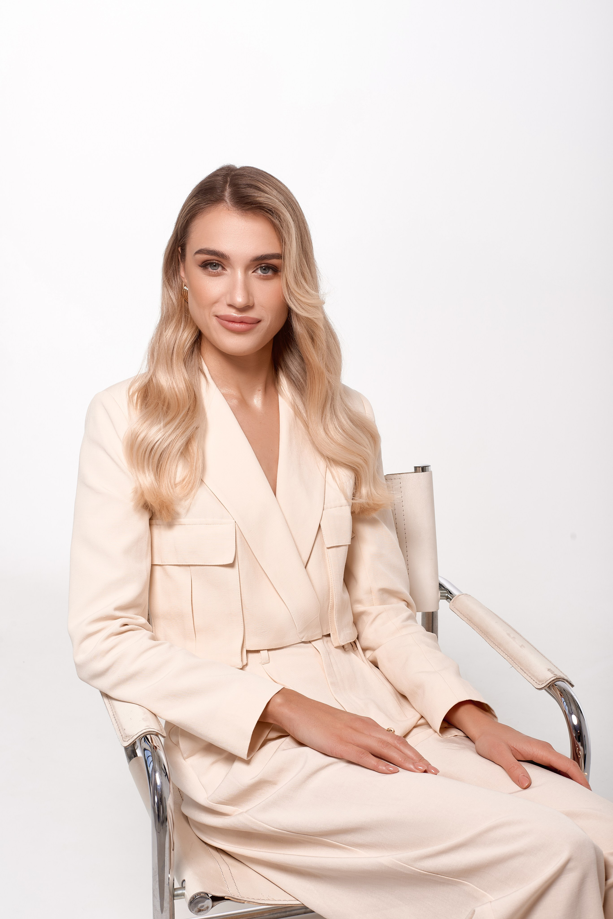Портретна студійна фотосесія блондинки у світлому брючному костюмі. Жінка сидить на кріслі, дивлячись в камеру з легкою усмішкою. Мінімалістичний фон додає акценту на образ.