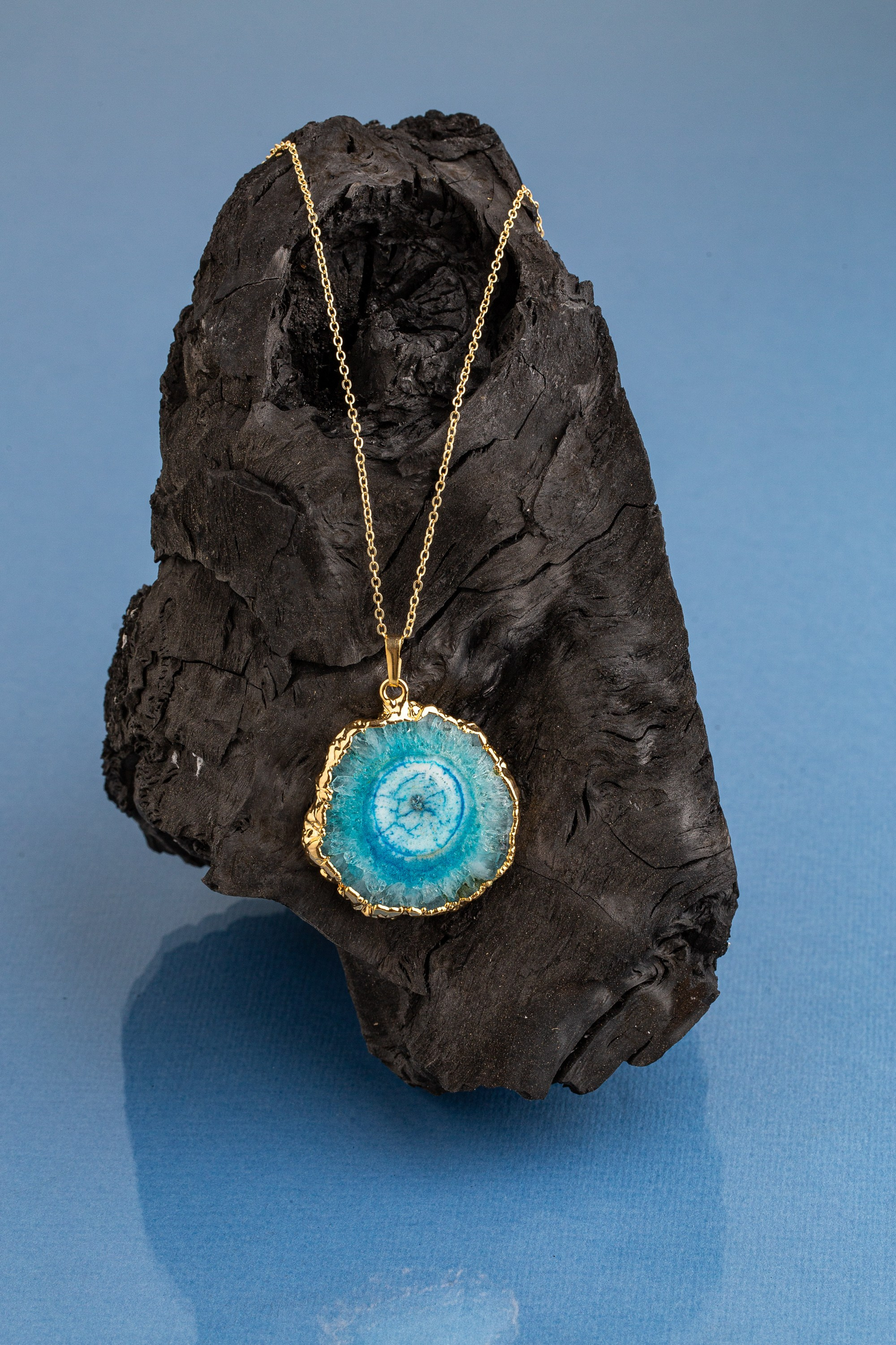 Tünd, teksturalı kömür və mavi fonda nümayiş etdirilən canlı mavi və ağ əqiq dilim kulonlu qızıl boyunbağı.