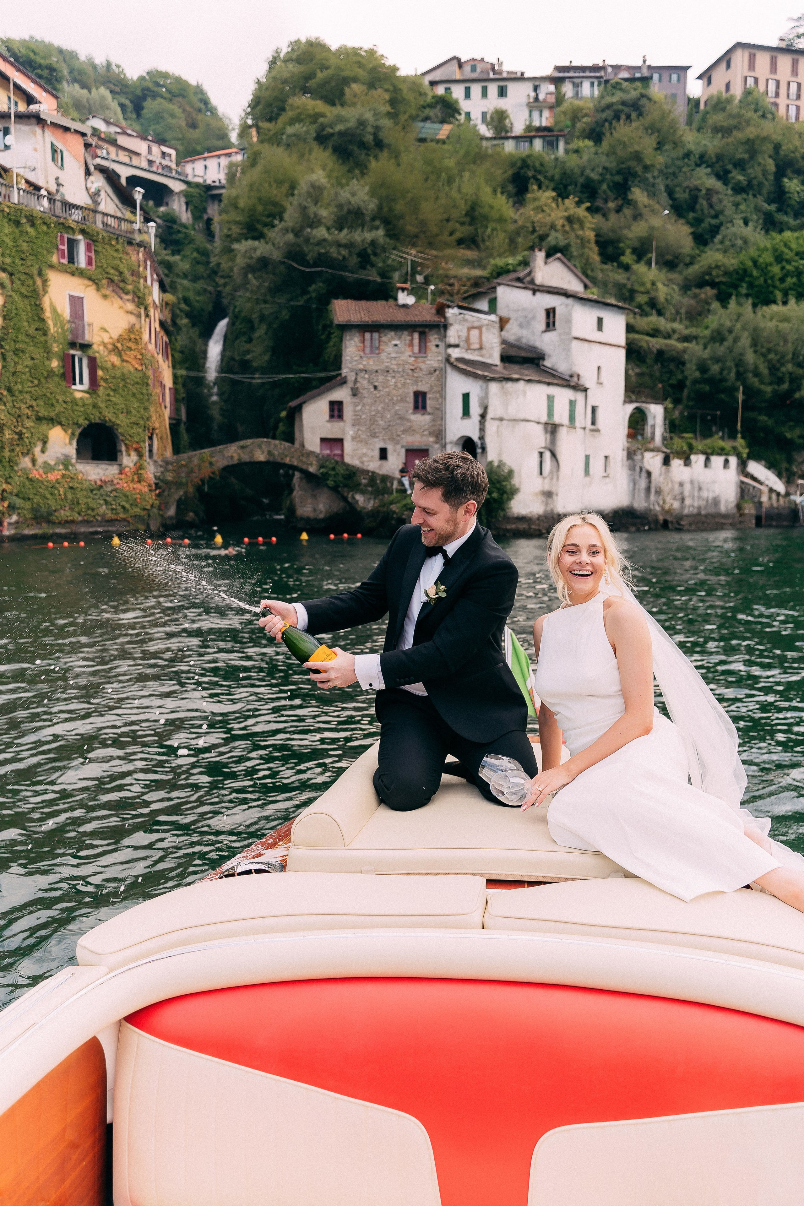 Bride and groom on the boat with the lakeside village at Villa Lario, Lake Como, Italy