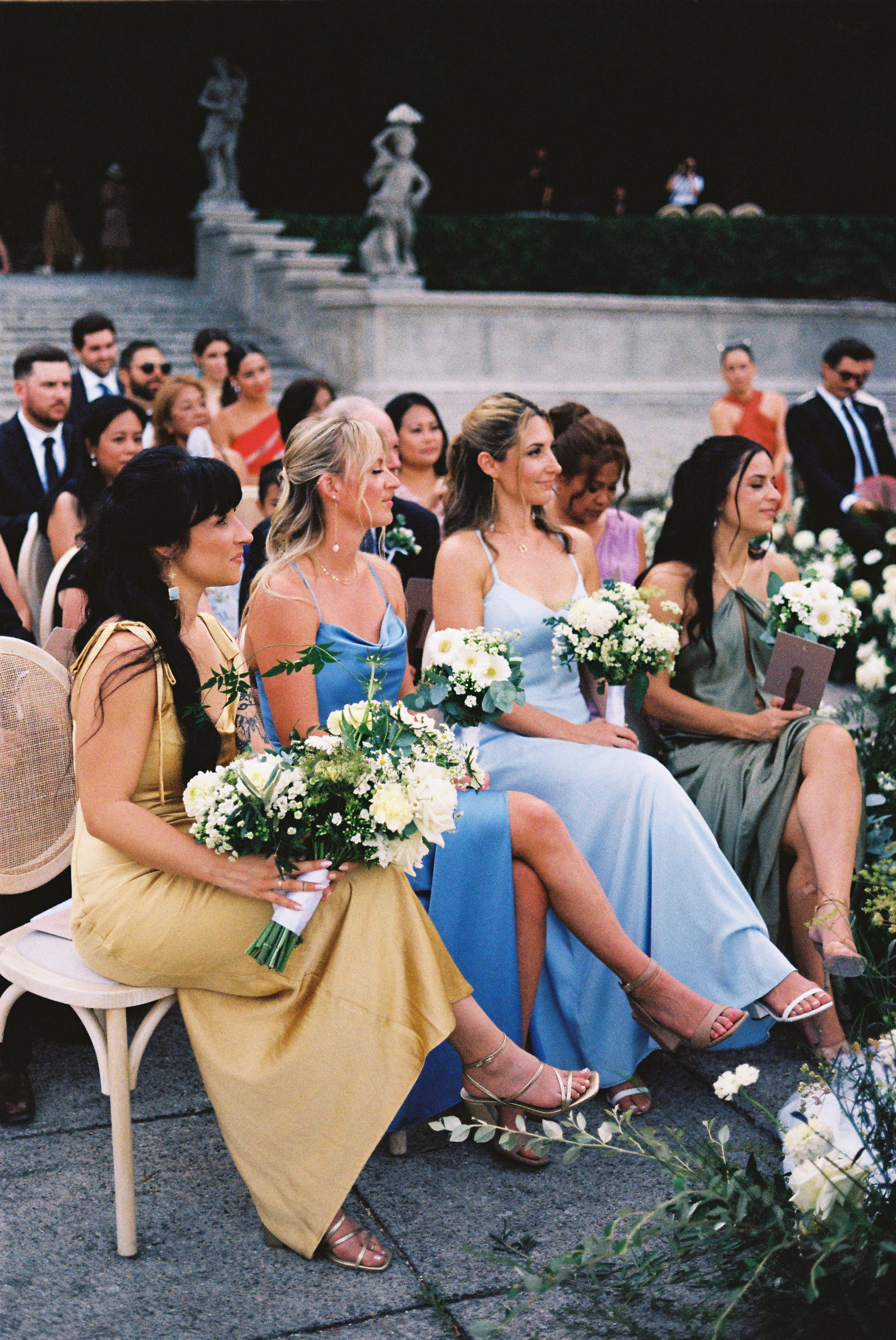 Bridesmaids in colorful dresses, holding white floral bouquets, attentively seated at wedding ceremony.