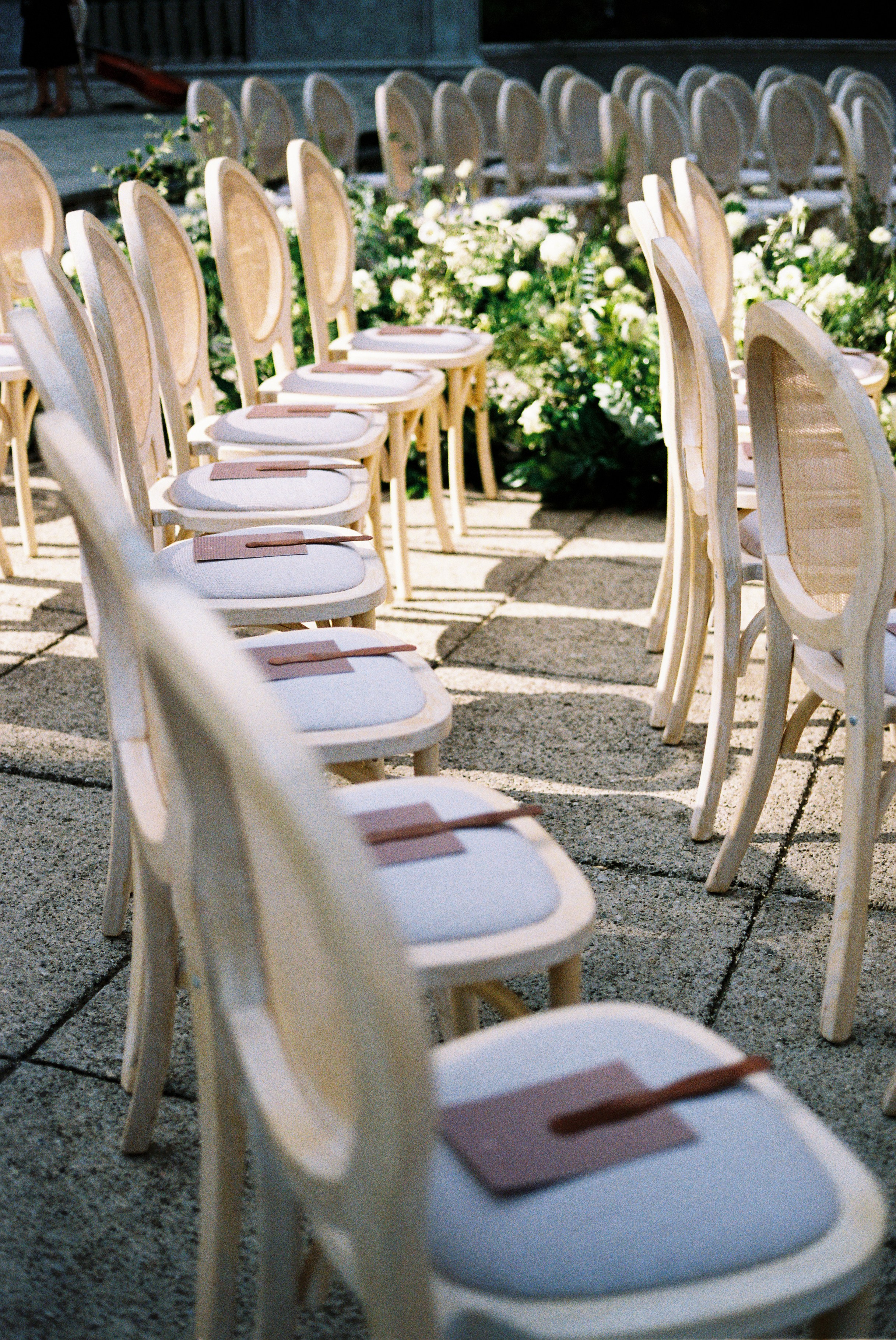 Rows of light-colored chairs with programs on seats, set for outdoor wedding ceremony.