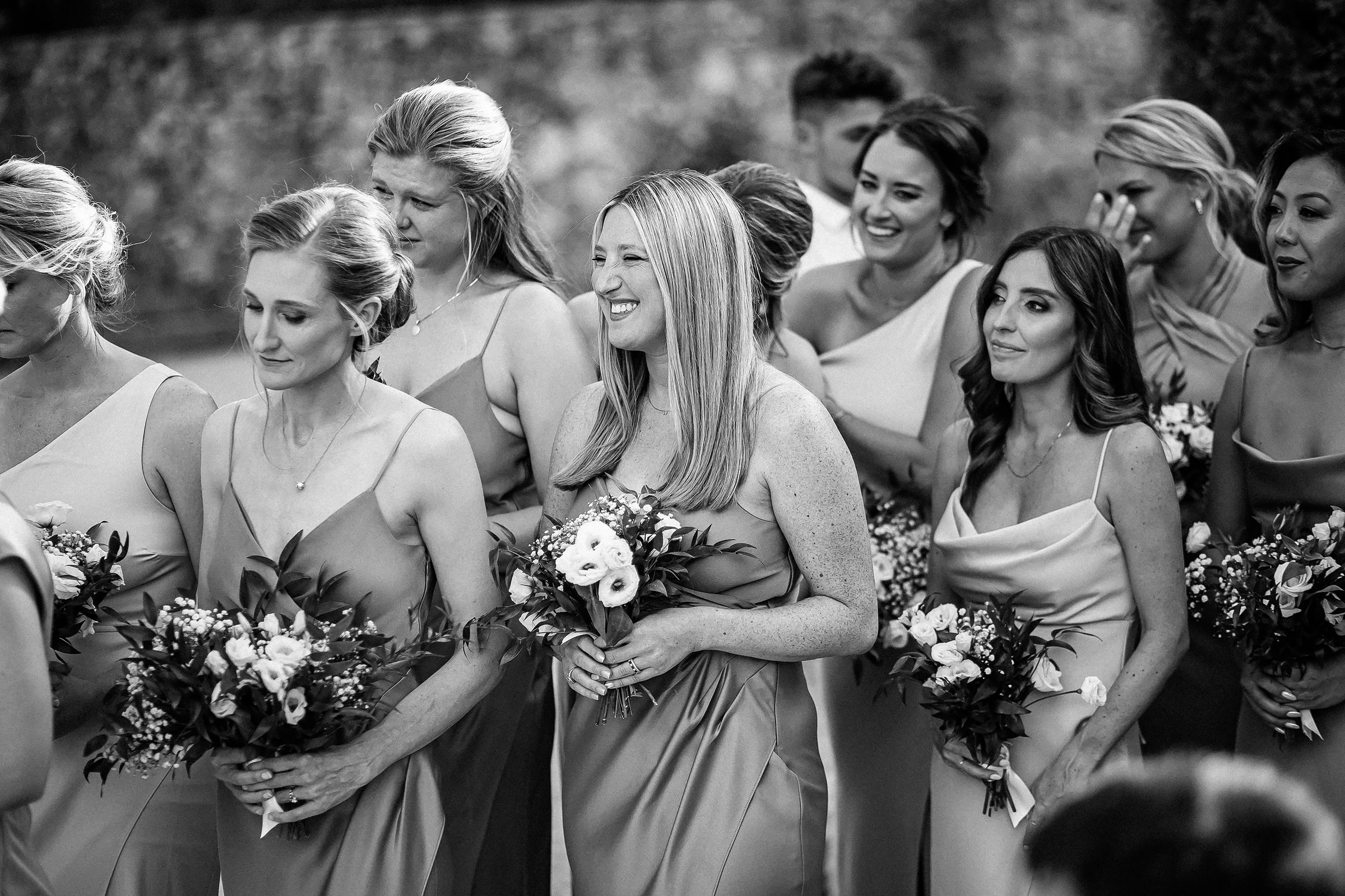 A group of bridesmaids in sage-green dresses, gathered together with radiant smiles, each holding beautifully arranged floral bouquets.