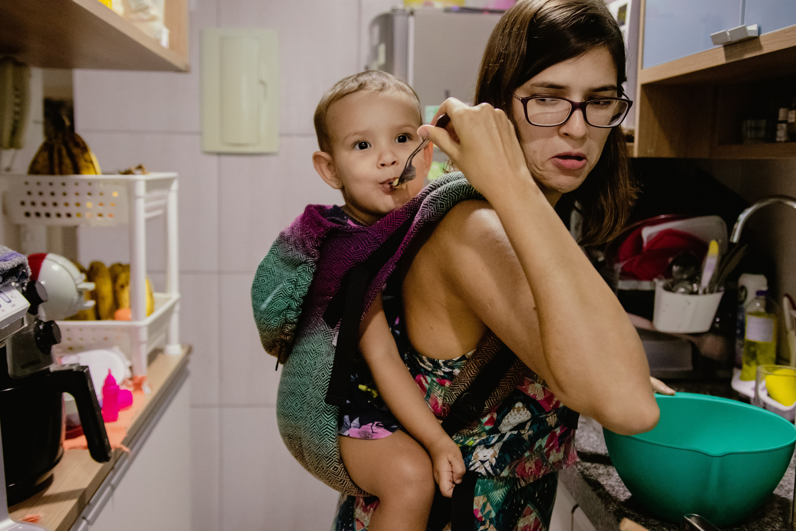 Mãe com filha no sling enquanto prepara comida em casa
