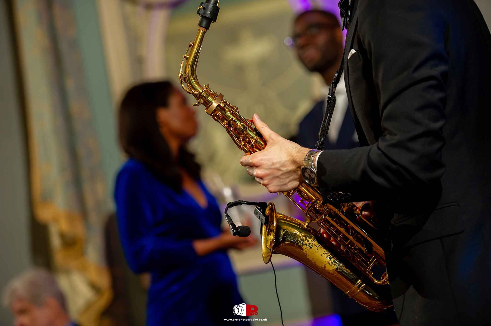A black tuxedo saxophonist performed live on stage during a private event, with guests blurred in the background.