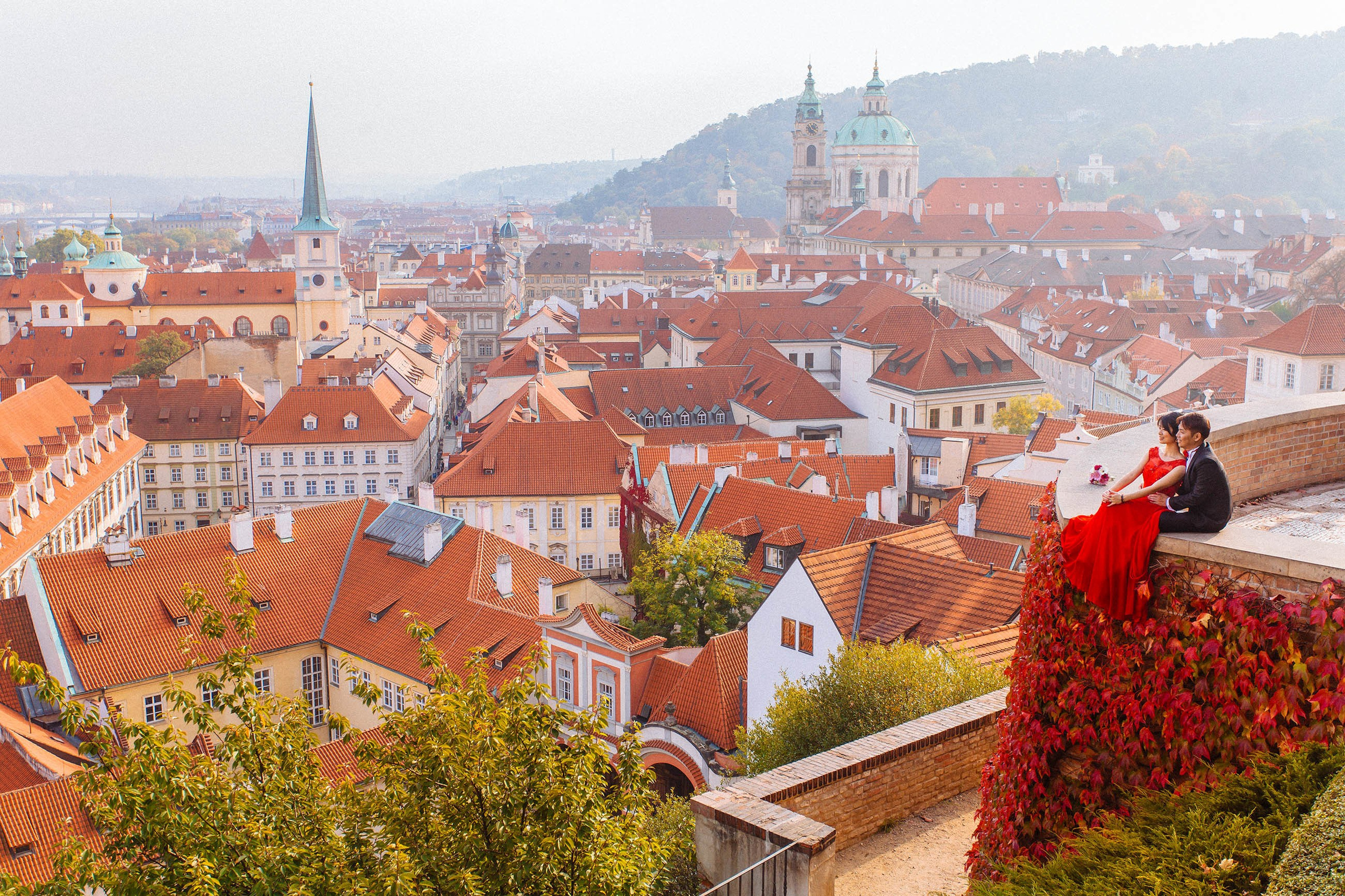 As the vibrant hues of Autum are seen among the red roofs of Prague, a red dress wearing Asian woman and her black-suited partner are seen sitting overlooking the city above at Prague Castle
