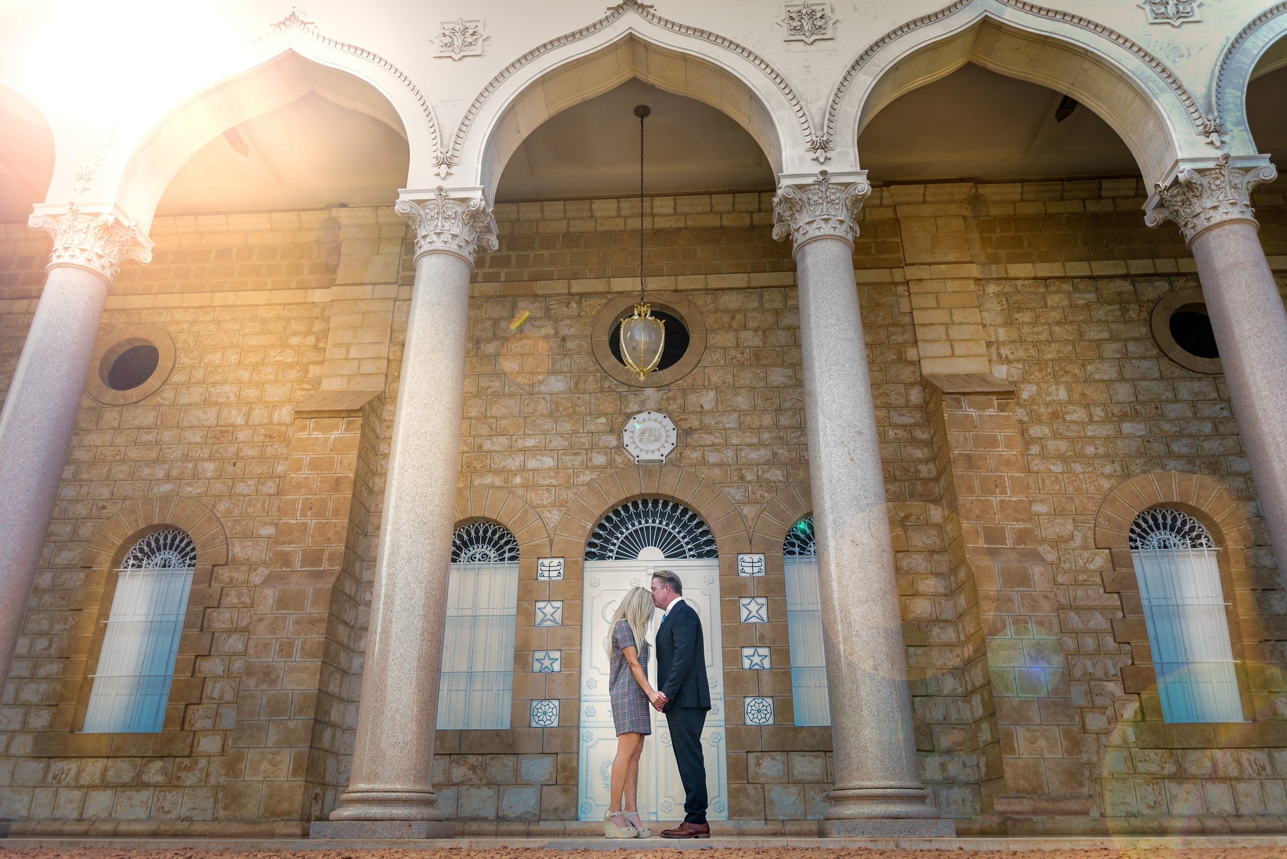 A couple stands in front of a majestic building with arches and columns in the Bahai Gardens in Haifa, enjoying a romantic photo shoot against the backdrop of historical architecture on a sunny day, holding hands