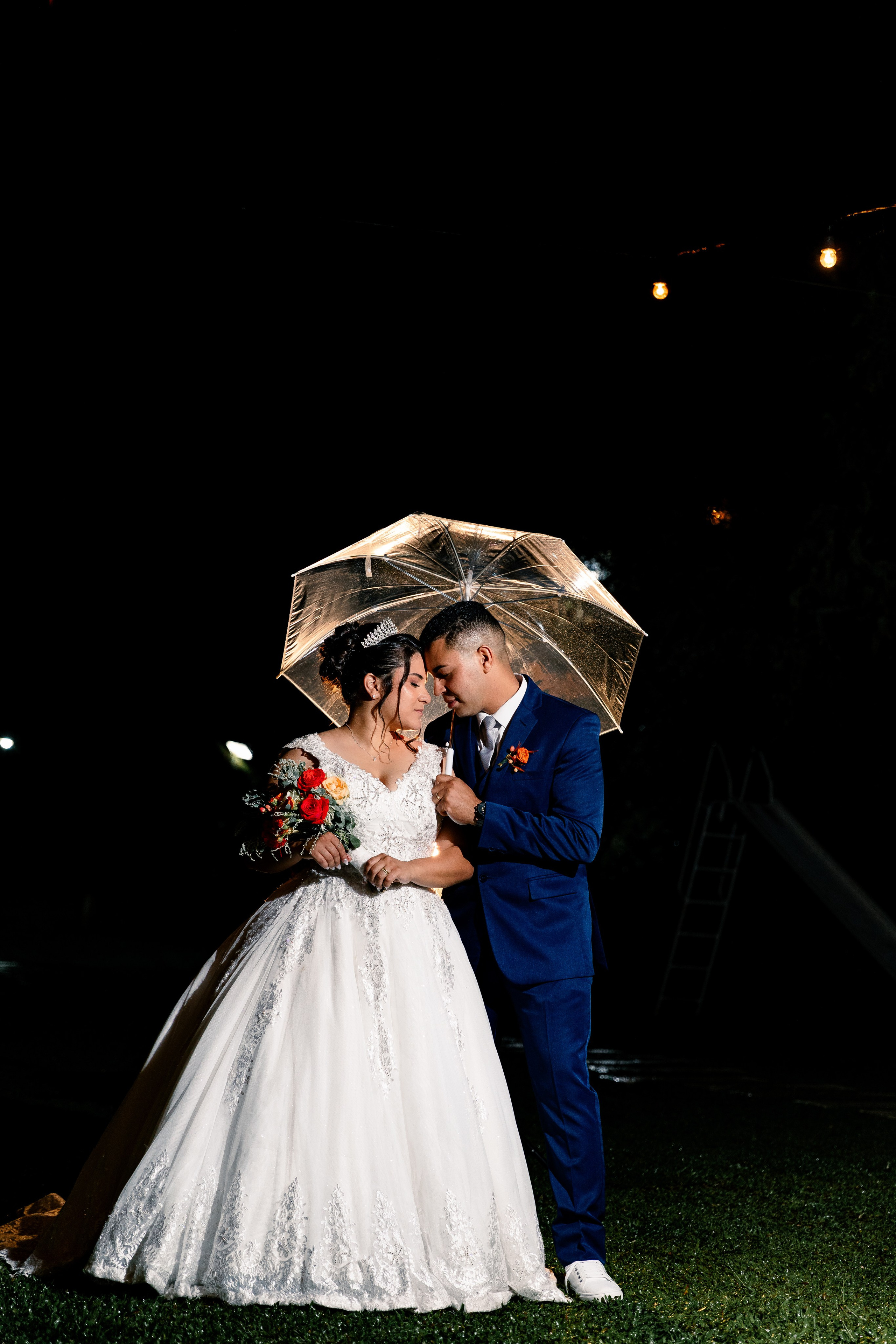 Noivos abraçados sob um guarda-chuva iluminado durante a noite, cena romântica e cinematográfica.