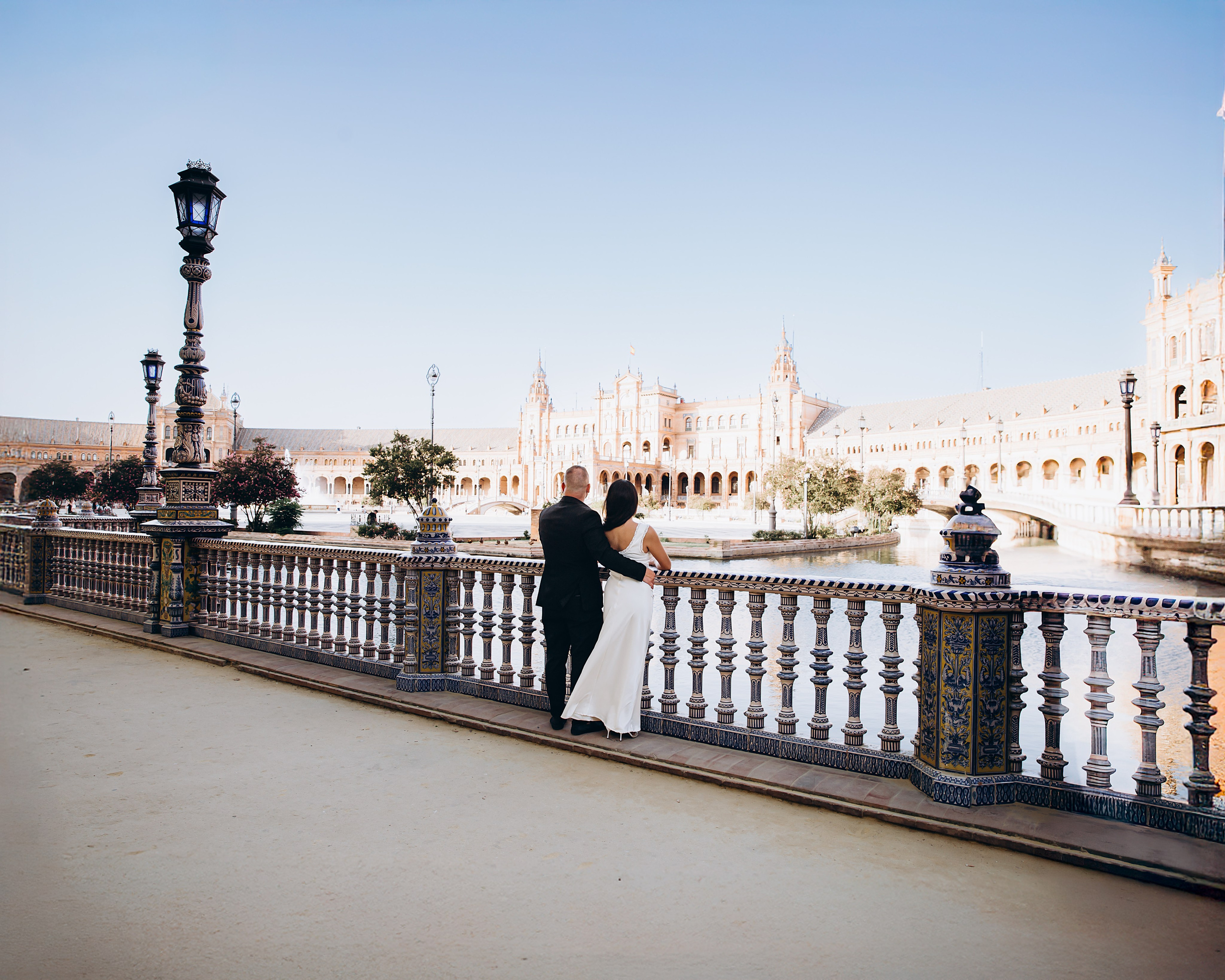 Couple embracing along the balustrade overlooking Plaza de España, Sevilla under a clear blue sky. Timeless scene from their civil wedding celebration in Andalusia.