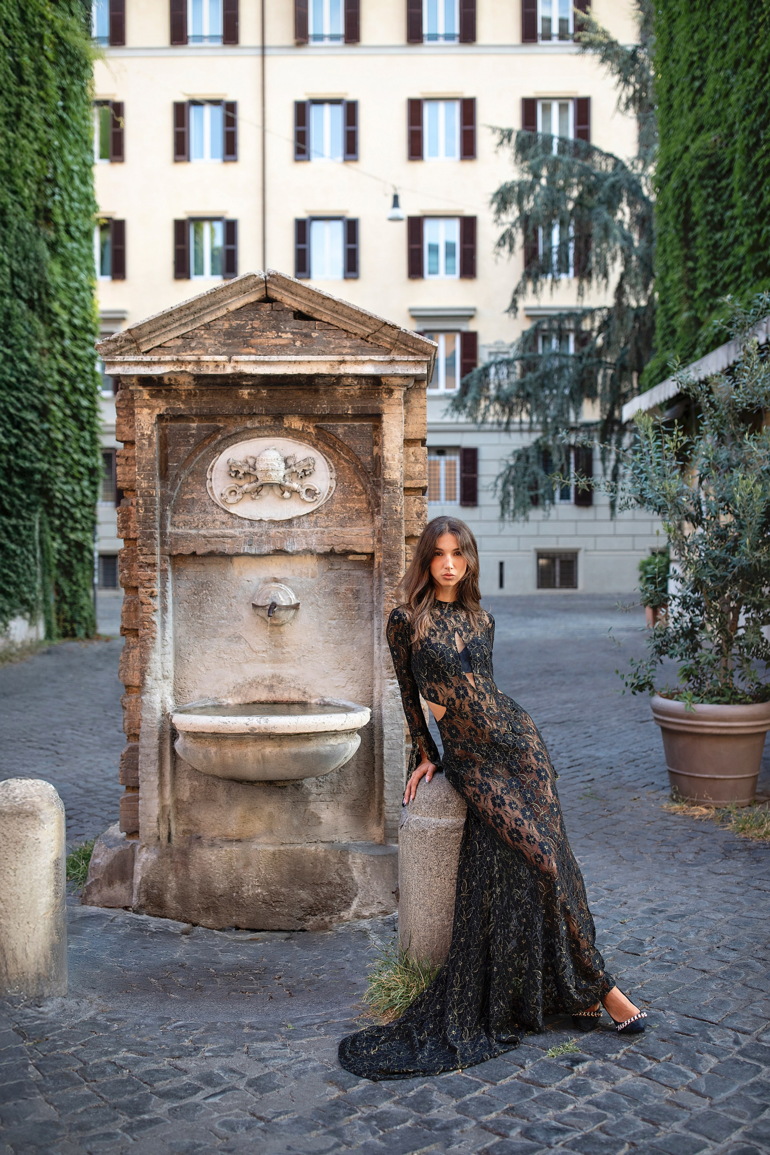 Elegant woman in a lace dress posing in front of an ancient Roman fountain during a fashion photoshoot in Rome.