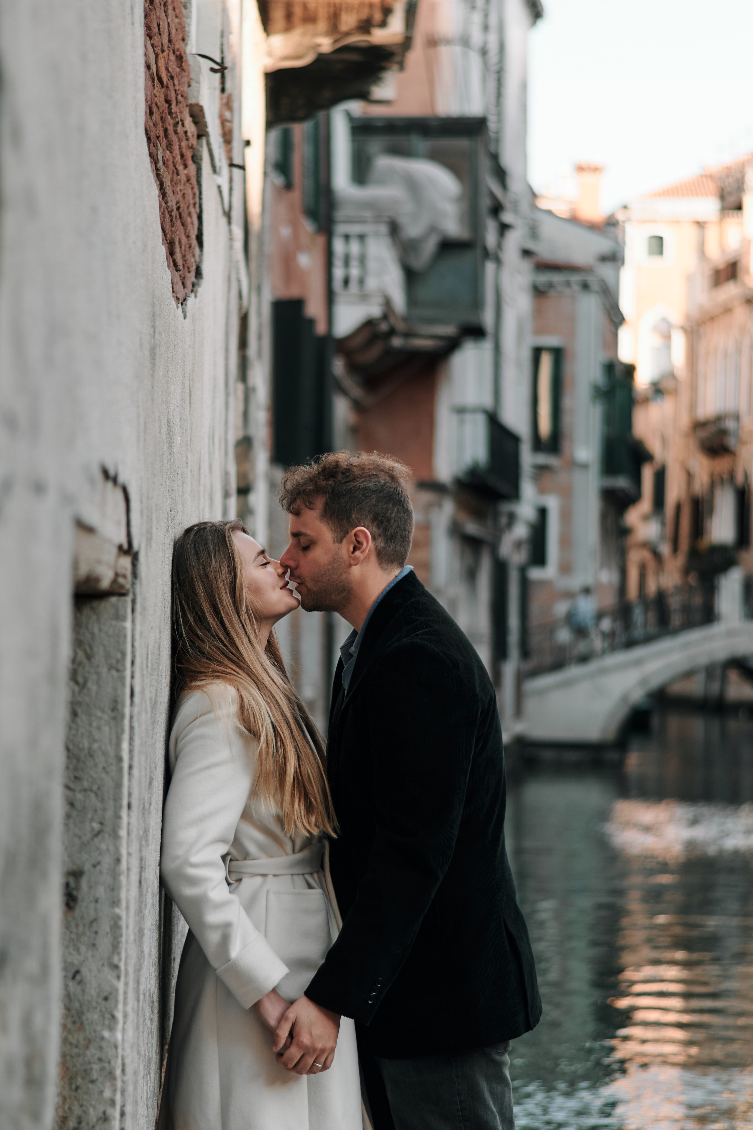 A romantic portrait captures a couple in the midst of a love story shoot on San Marco Square. They stand close together, gazing out at the iconic Bridge of Sighs in the distance. The photographer has expertly framed the shot to showcase both the couple's love and the beauty of Venice's architecture.