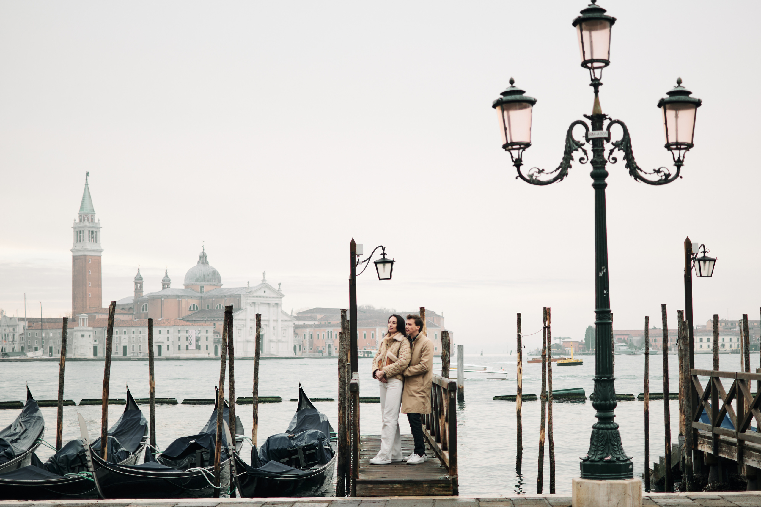 Against the stunning backdrop of San Marco Square, a couple stands hand in hand, their love for each other evident in their smiles and the way they look at each other. The Columns of San Marco and San Todaro frame the couple, adding to the drama and beauty of the photo.