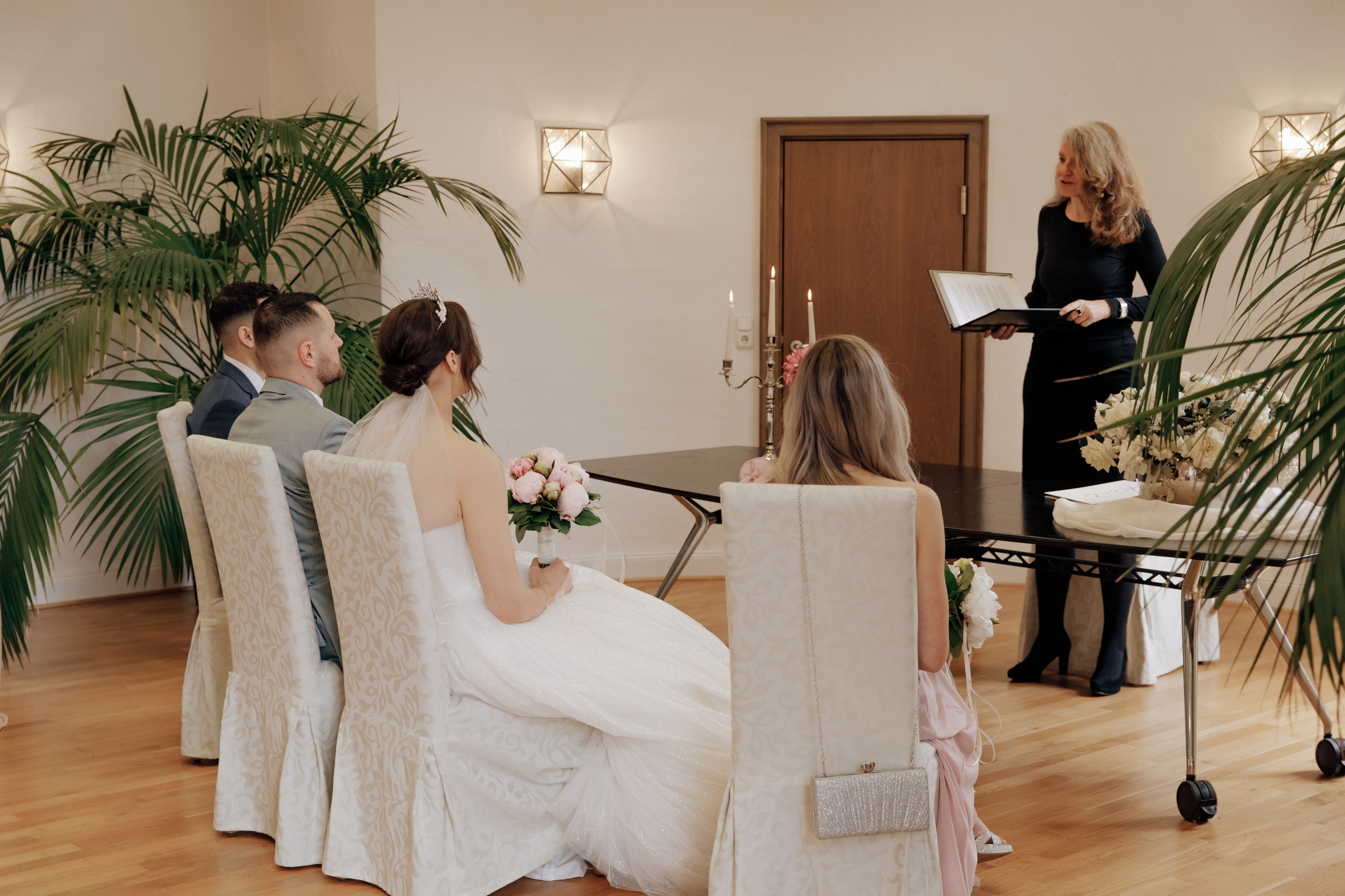 Die Standesbeamte hält eine Rede, während der Bräutigam, die Braut und die Trauzeugen mit dem Rücken zur Kamera zuhören. 