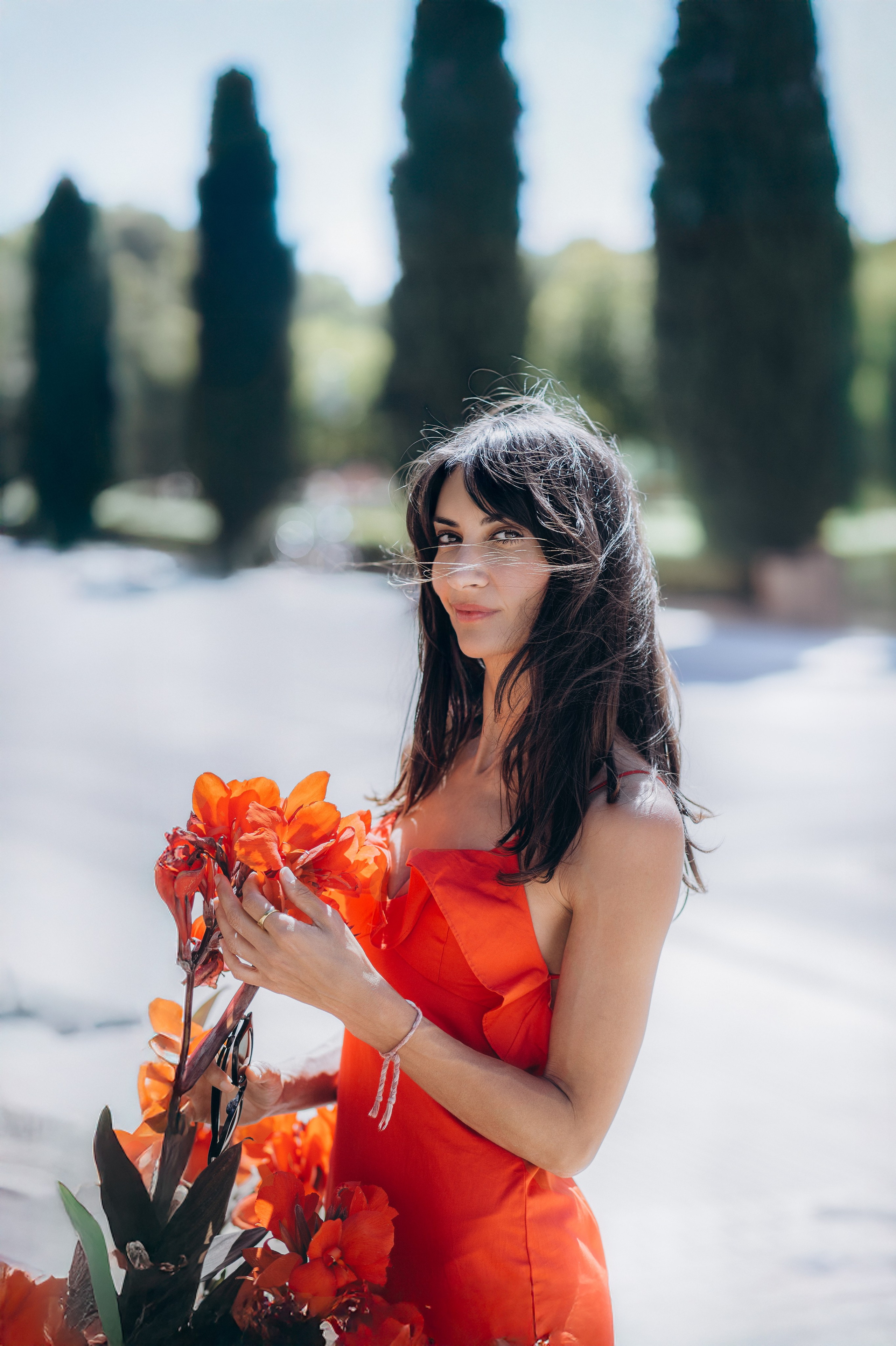 Элегантная женщина в ярко-красном платье с букетом тропических оранжевых цветов, стоящая под мягким естественным светом на фоне кипарисов — художественная портретная фотография в Кальпе, Испания, передающая цвет, красоту и эмоции в спокойной обстановке на открытом воздухе.