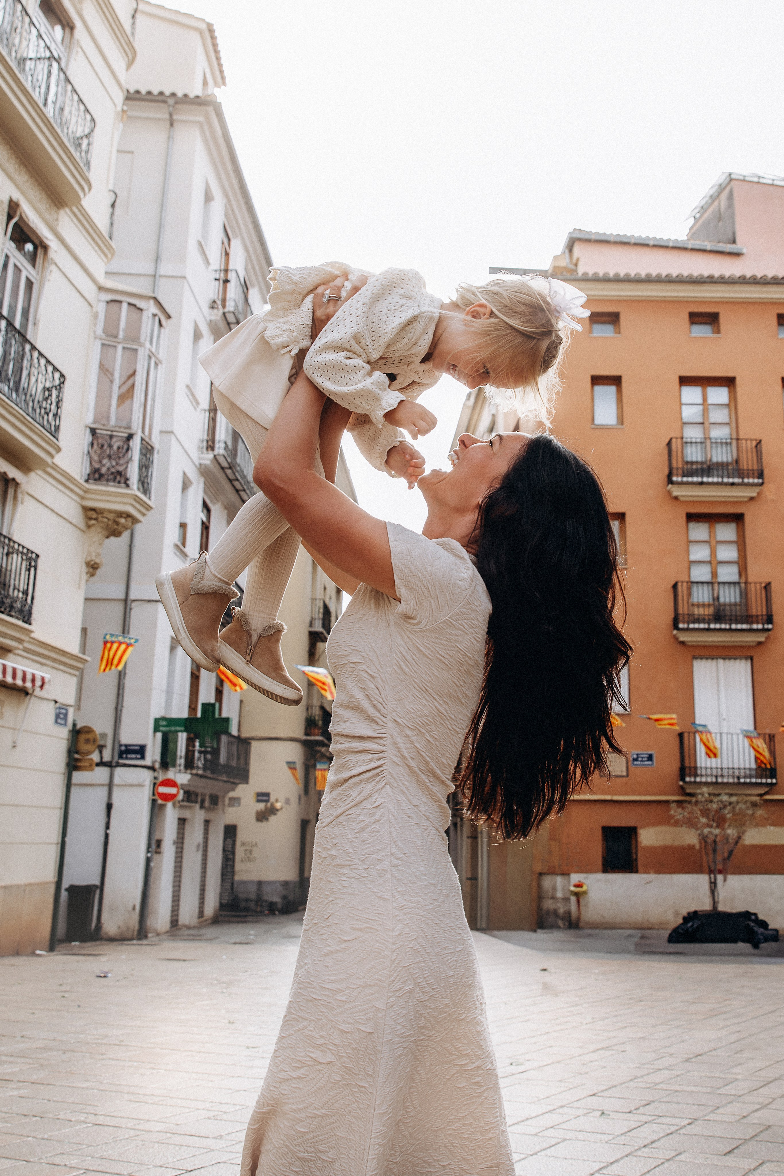 Sesión de fotos familiar en Valencia, España, con una madre levantando con alegría a su pequeña hija en una histórica plaza del casco antiguo rodeada de la hermosa arquitectura mediterránea. Un emotivo retrato lifestyle de madre e hija que transmite risas, amor y momentos familiares inolvidables. Ideal para fotografía familiar, sesiones de fotos familiares, retratos de madre e hija, fotografía infantil, fotografía de vacaciones y fotografía familiar de destino en Valencia y toda España.