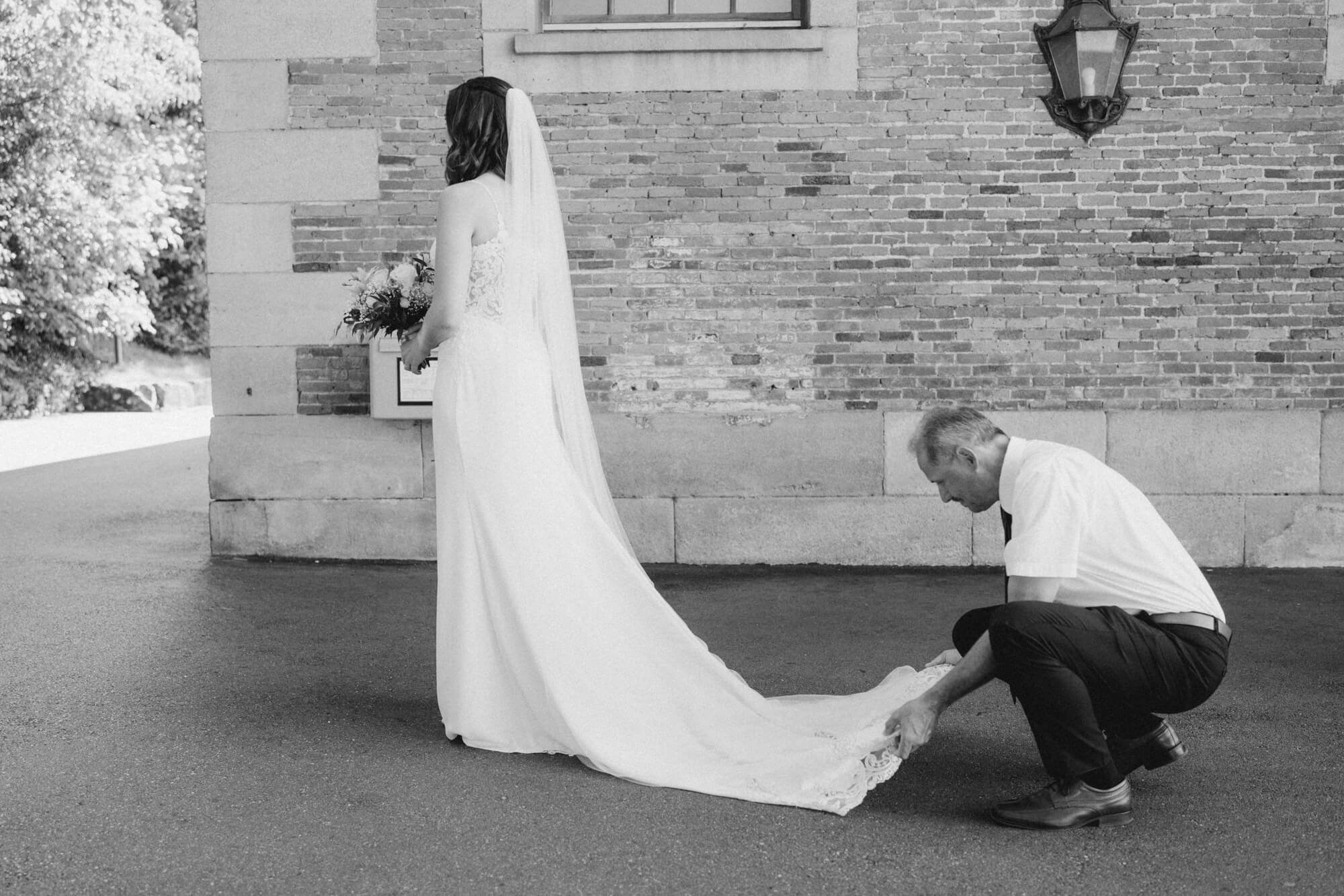 Guest kneeling to arrange the bride's dress train outside a sandstone building in Stuttgart; black and white