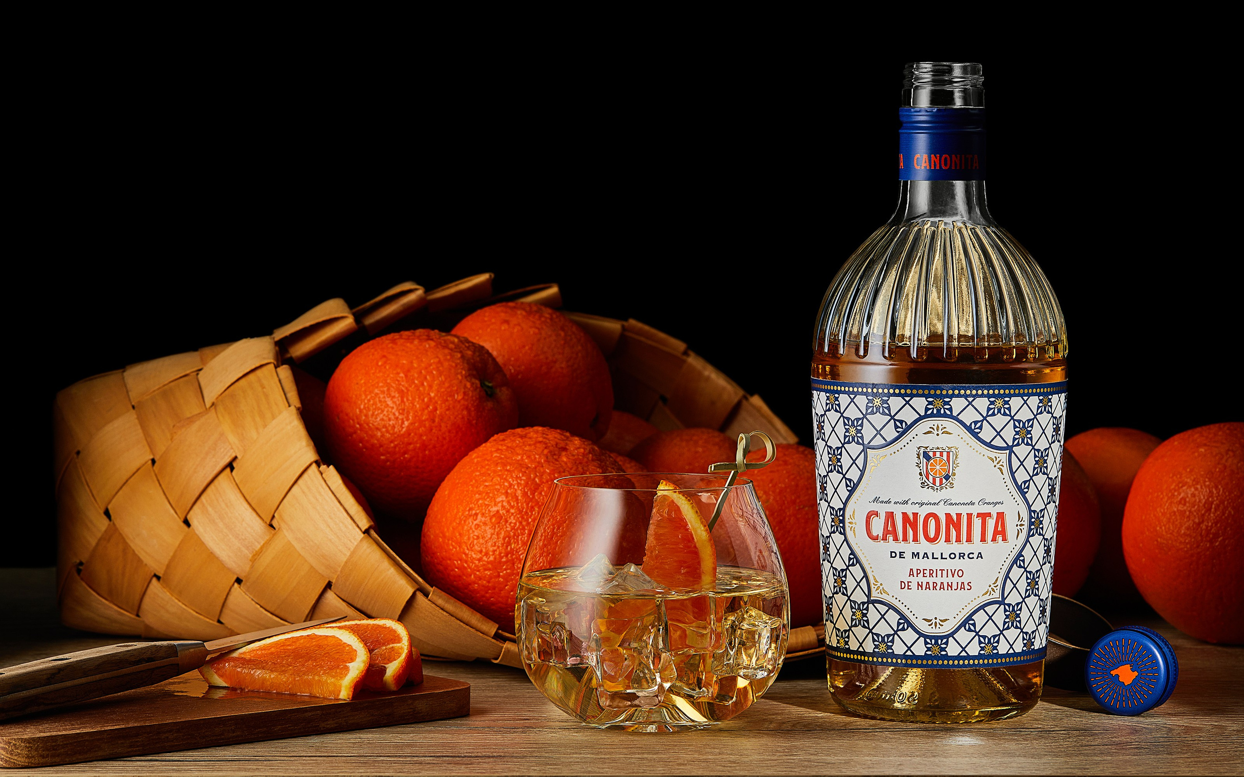 Photographer Roman Djuzev - The photograph features a bottle of Canonita de Mallorca aperitif with a striking decorative label in a Mediterranean style. The golden-hued drink is beautifully lit, highlighting the textured shape of the bottle. Beside it stands a low glass tumbler with ice and an orange slice, creating the impression of a fresh, chilled cocktail.