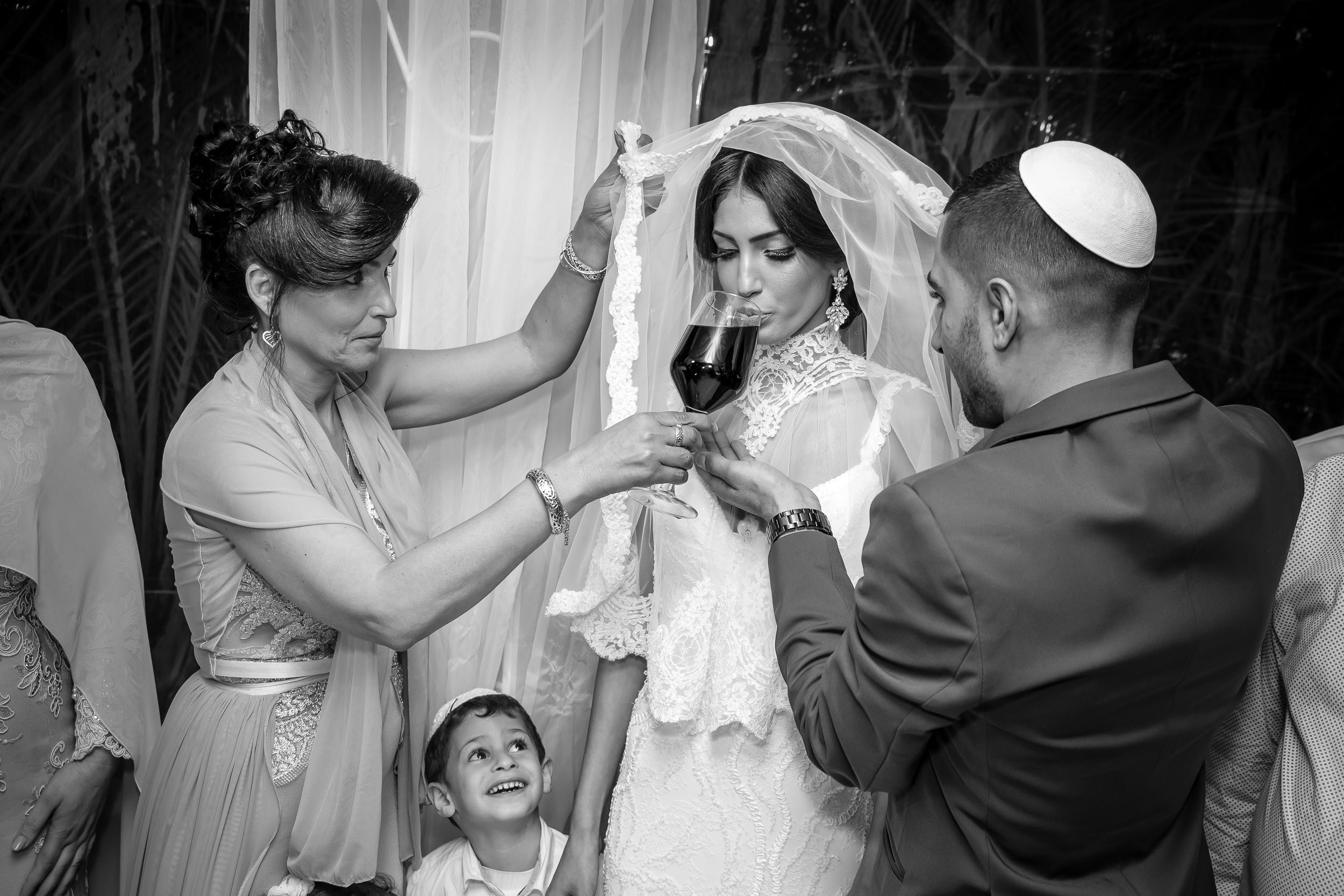 A bride drinks from a wine glass held by the groom beneath the chuppah