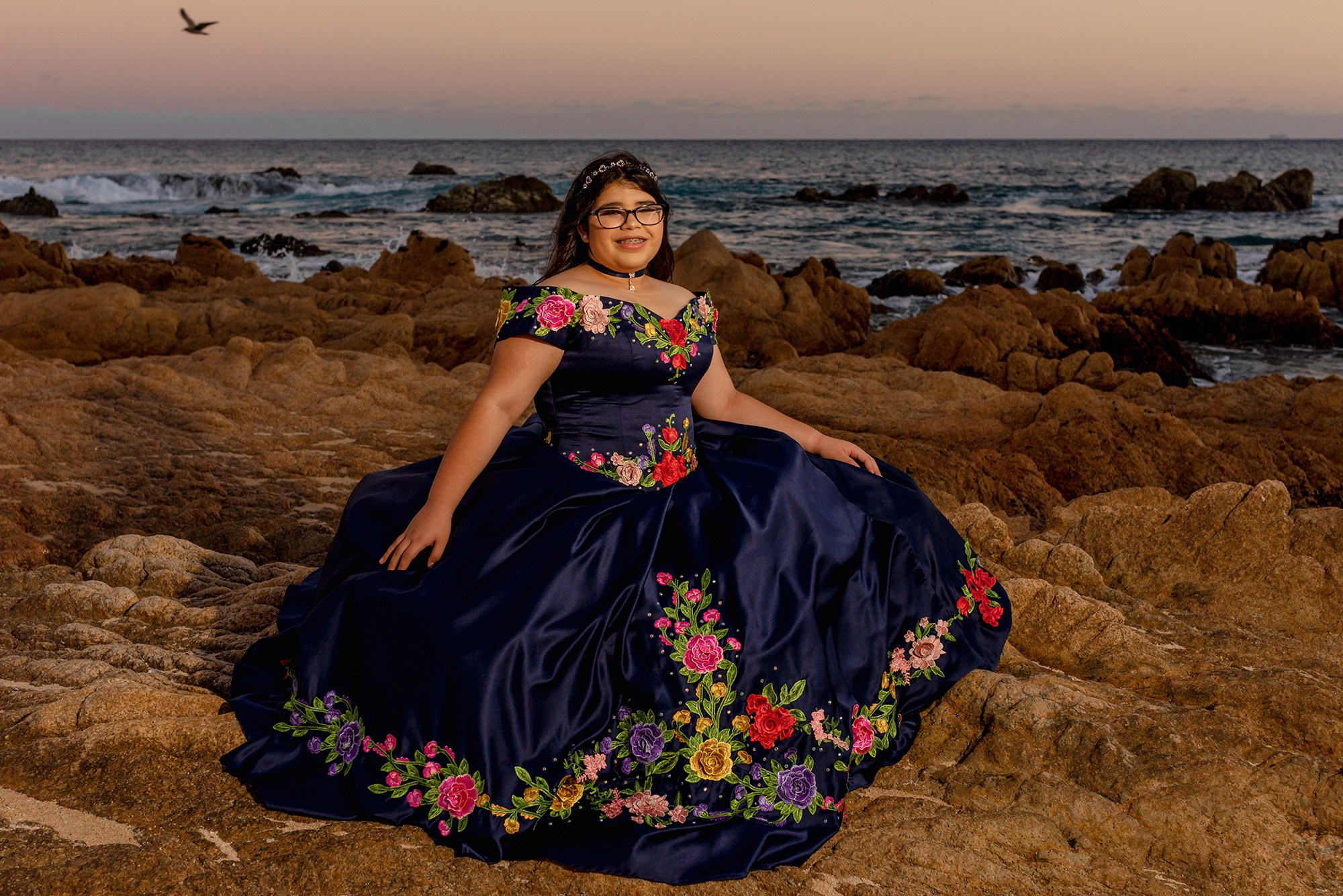 Quinceañera photography in Los Cabos – girl in a traditional Mexican blue dress with floral embroidery, holding a multicolor bouquet during sunset session at Las Viudas beach